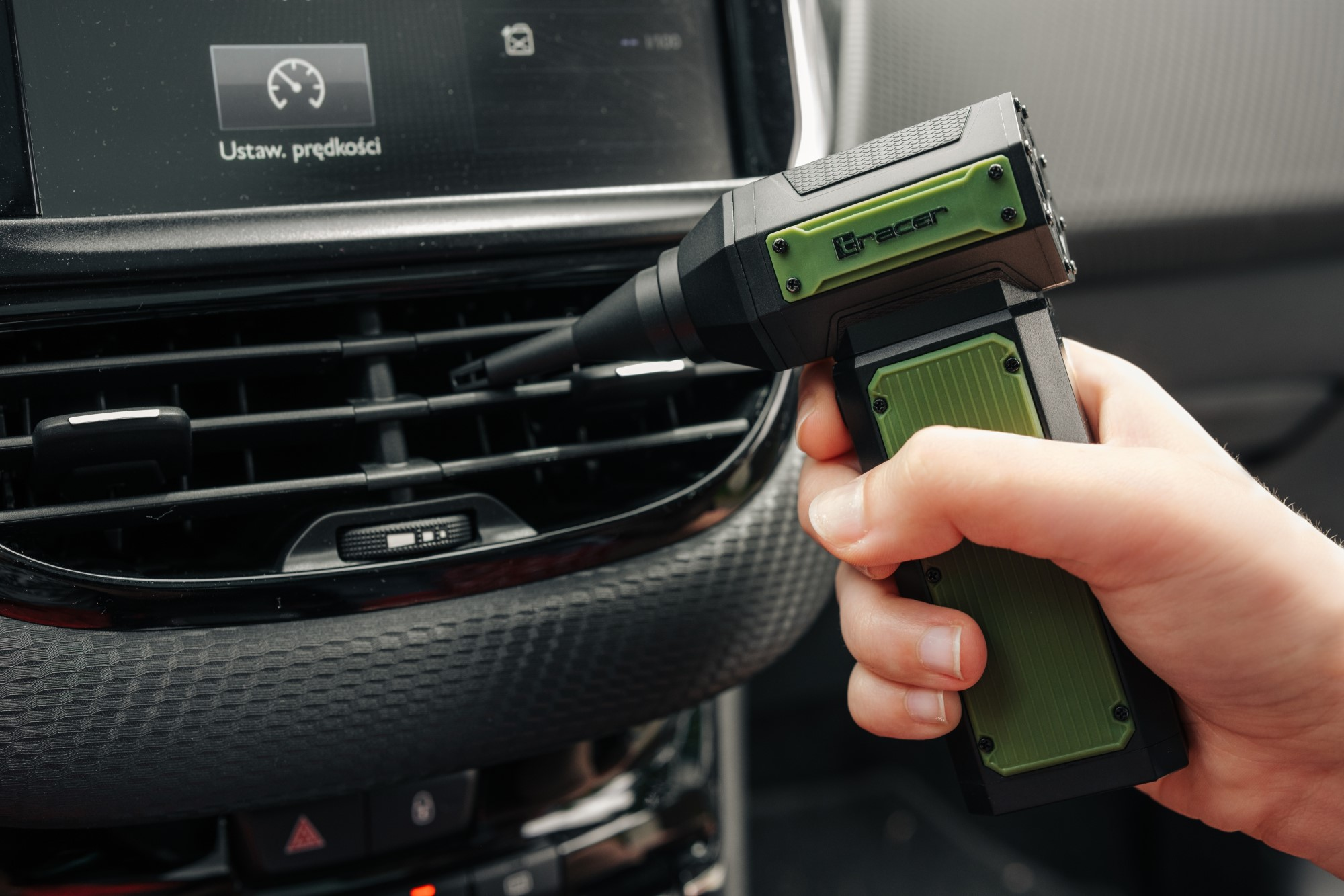 Ręka trzymająca zielono-czarne urządzenie Tracer, kierująca dyszę na kratkę wentylacyjną samochodu. Widoczna deska rozdzielcza.