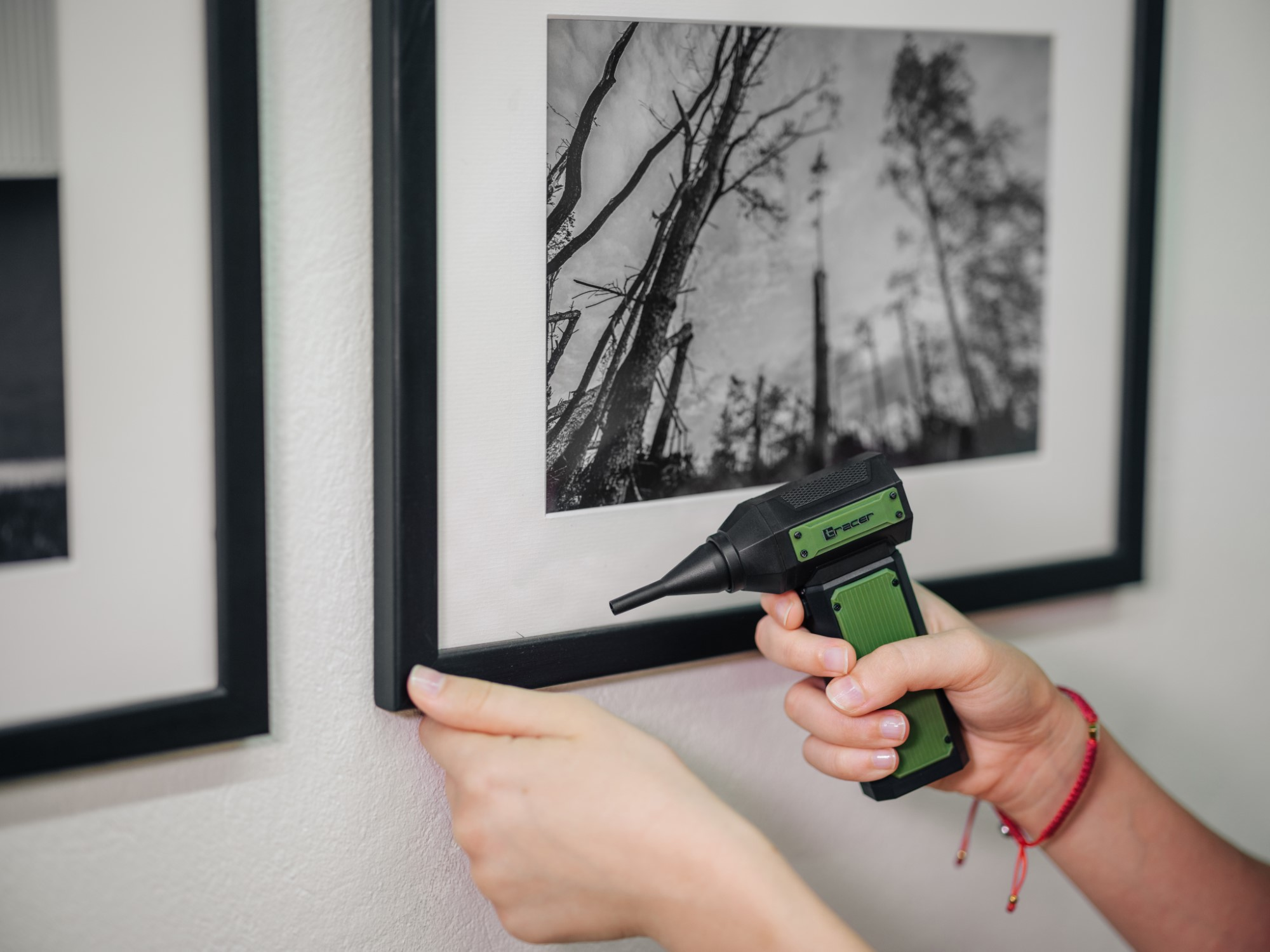 Osoba trzymająca zielono-czarne narzędzie, dopasowująca oprawione zdjęcie do białej ściany. Widoczne jest kolejne oprawione zdjęcie.