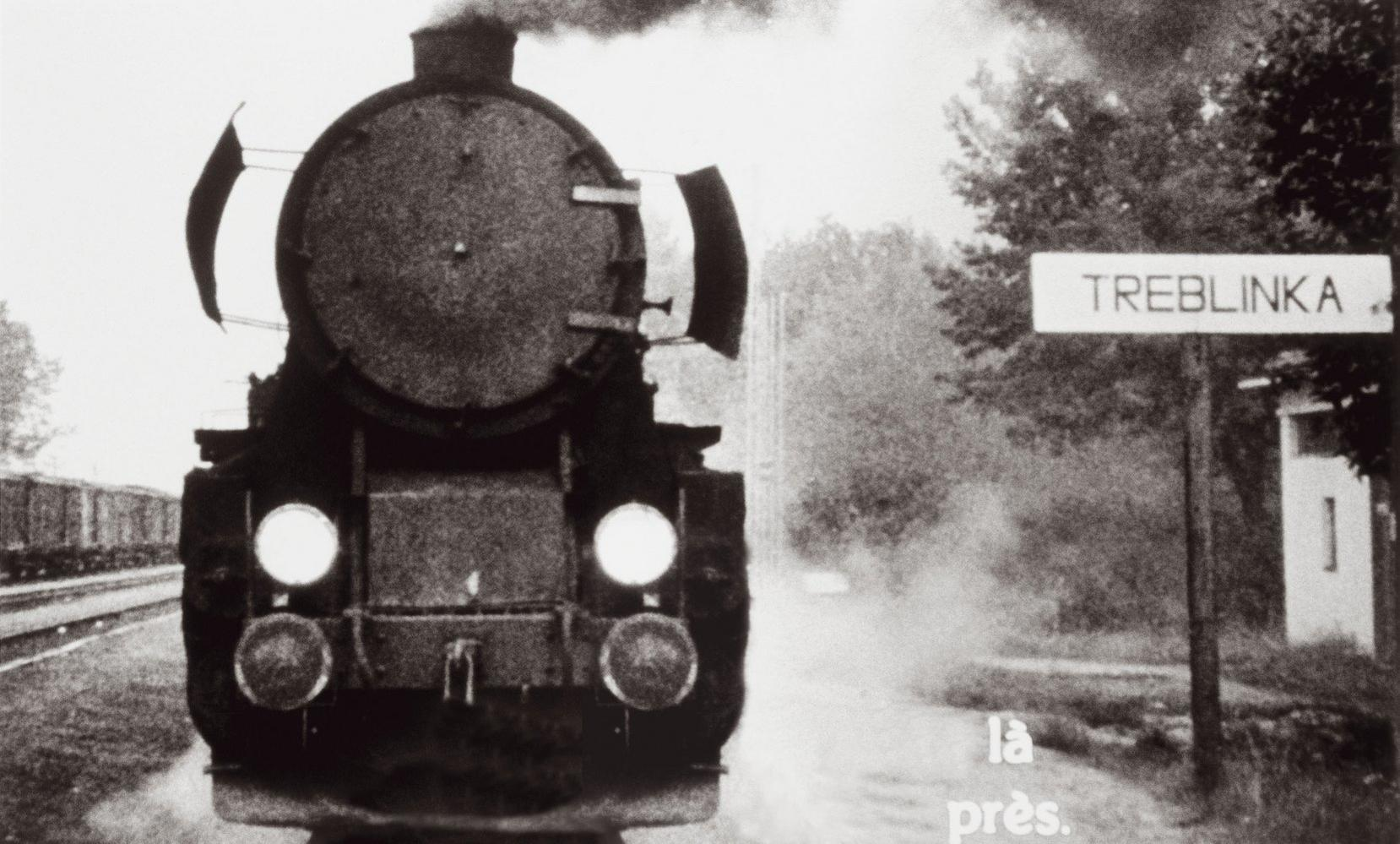 Schwarz-Weiß-Bild eines Dampfzuges am Bahnhof Treblinka. Rauch quillt aus dem Schornstein. Ein Schild trägt die Aufschrift 'Treblinka'.