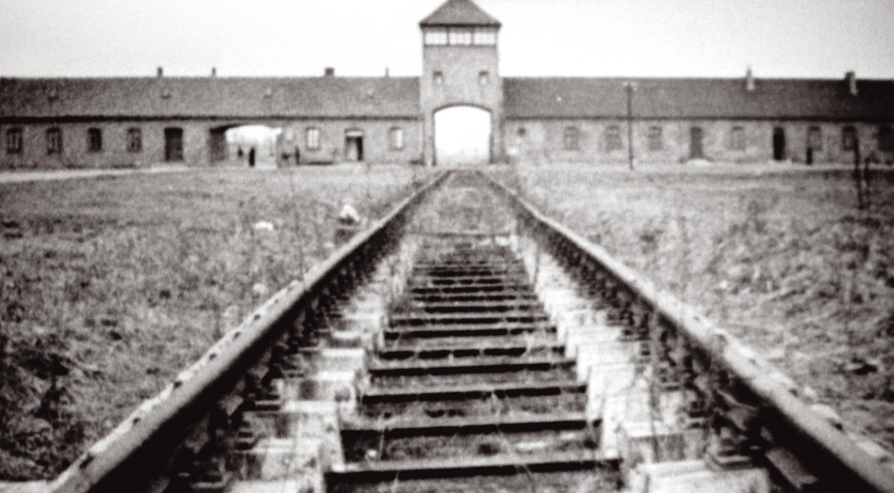 Schwarz-Weiß-Bild des Auschwitz-Tors mit Bahngleisen, die darauf zuführen. Gebäude auf beiden Seiten.