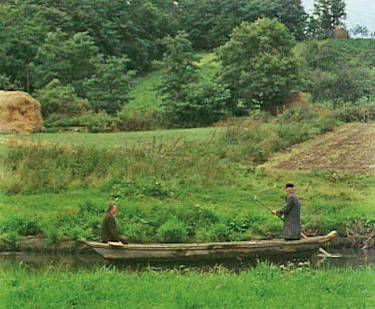 Zwei Männer in einem Holzboot auf einem Fluss. Grüne Felder und Bäume im Hintergrund. Ein Mann rudert mit einer Stange.