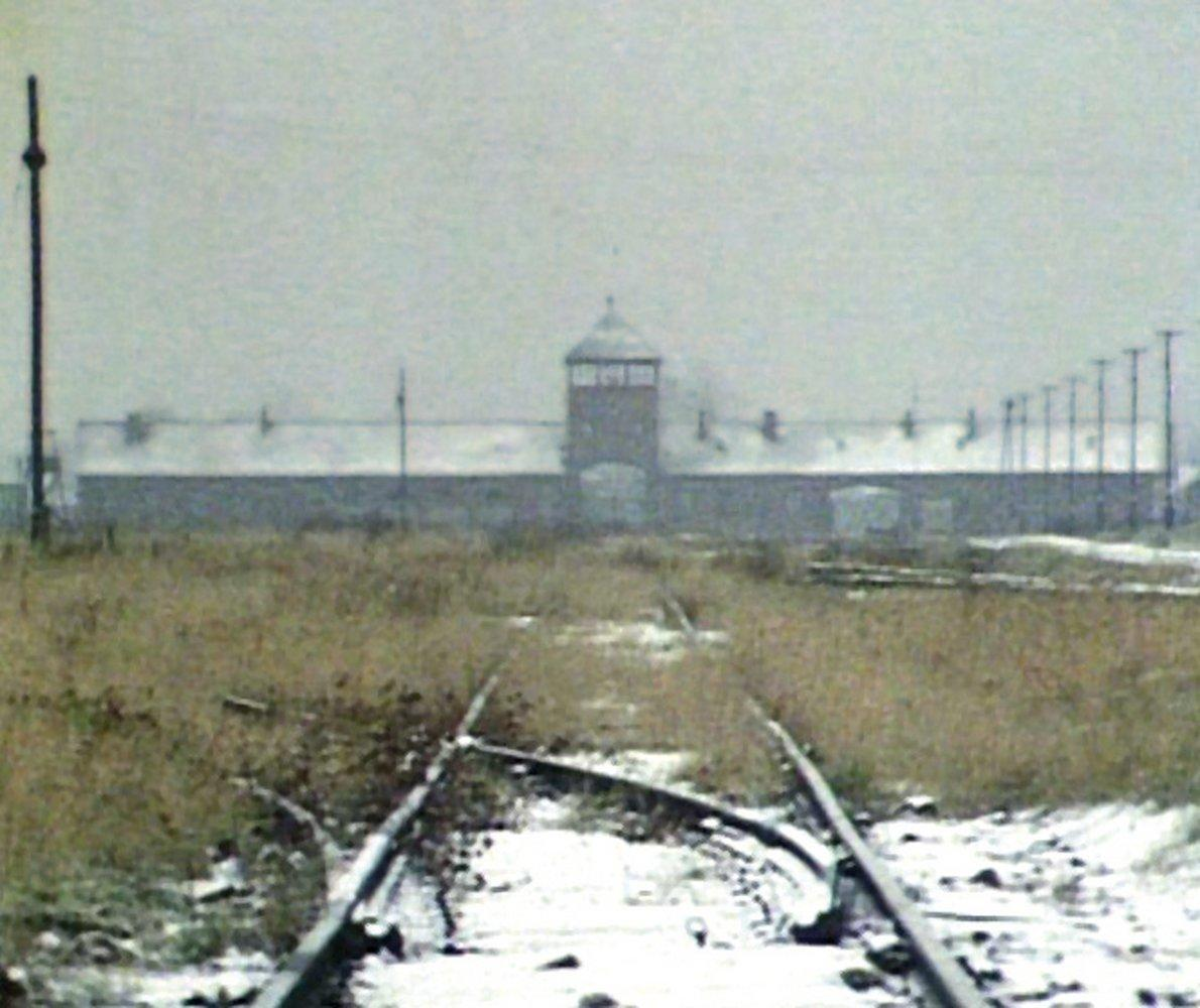 Bahngleise führen zu einem Gebäude mit Turm. Die Szene ist verschneit, mit Gras und Masten. Der Himmel ist bedeckt.