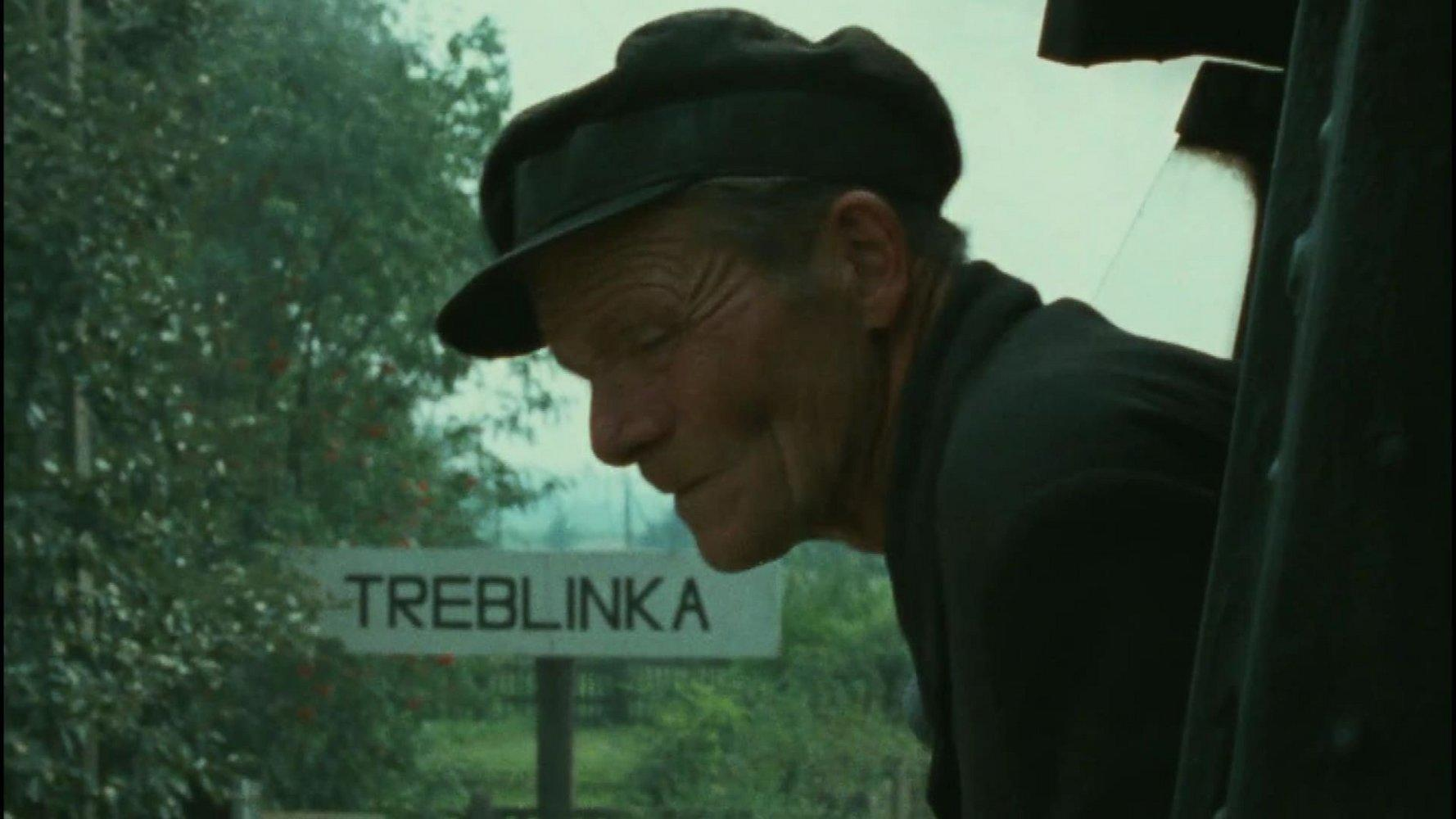 Ein älterer Mann mit Mütze blickt nach unten. Ein Schild zeigt Treblinka. Grüne Bäume im Hintergrund. Die Szene ist wahrscheinlich historisch.