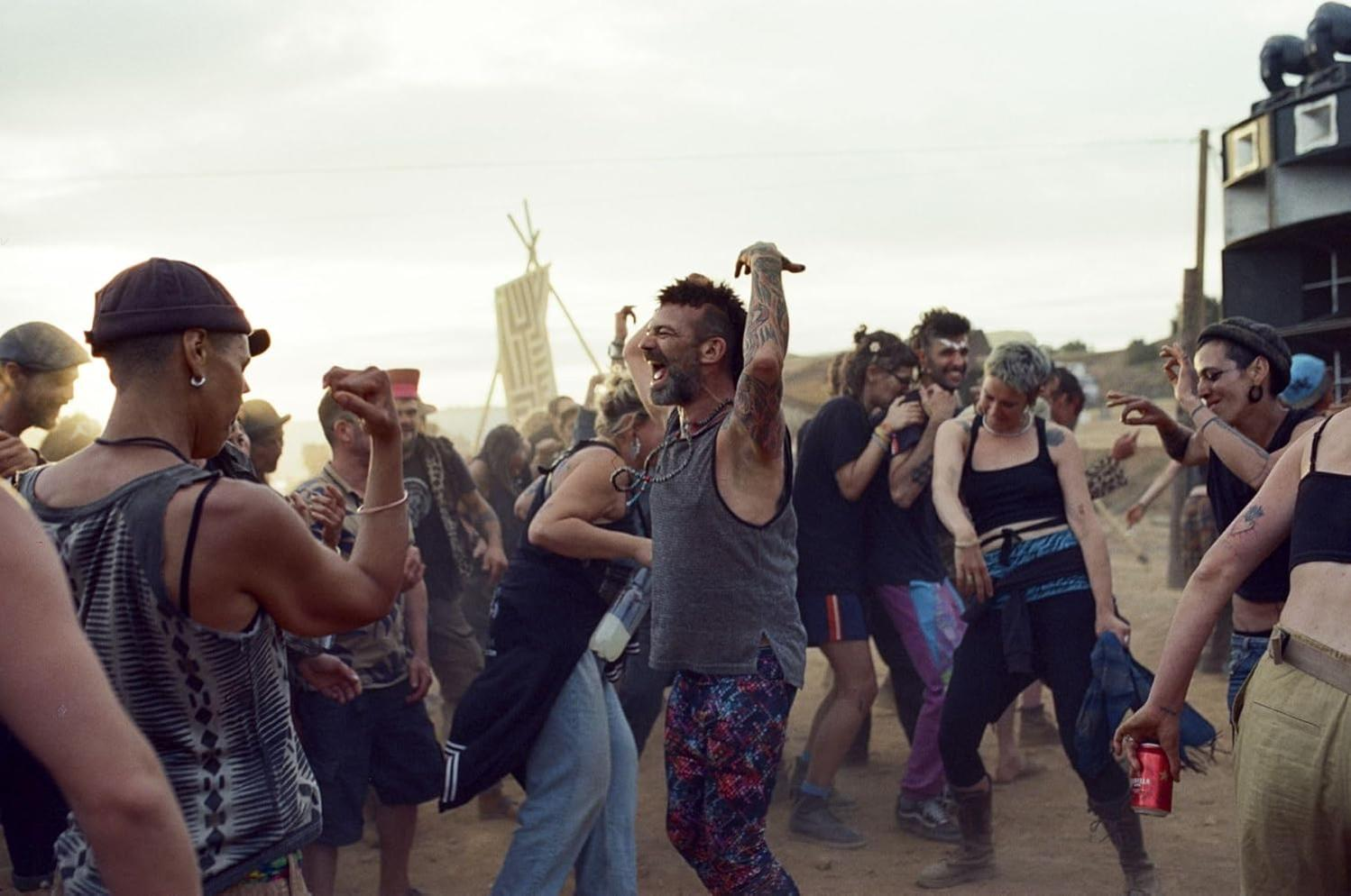Menschen tanzen bei einer Outdoor-Veranstaltung. Ein Mann mit Tattoos hebt die Arme. Andere lächeln, einige halten Getränke. Ein Lautsprecherturm ist sichtbar.