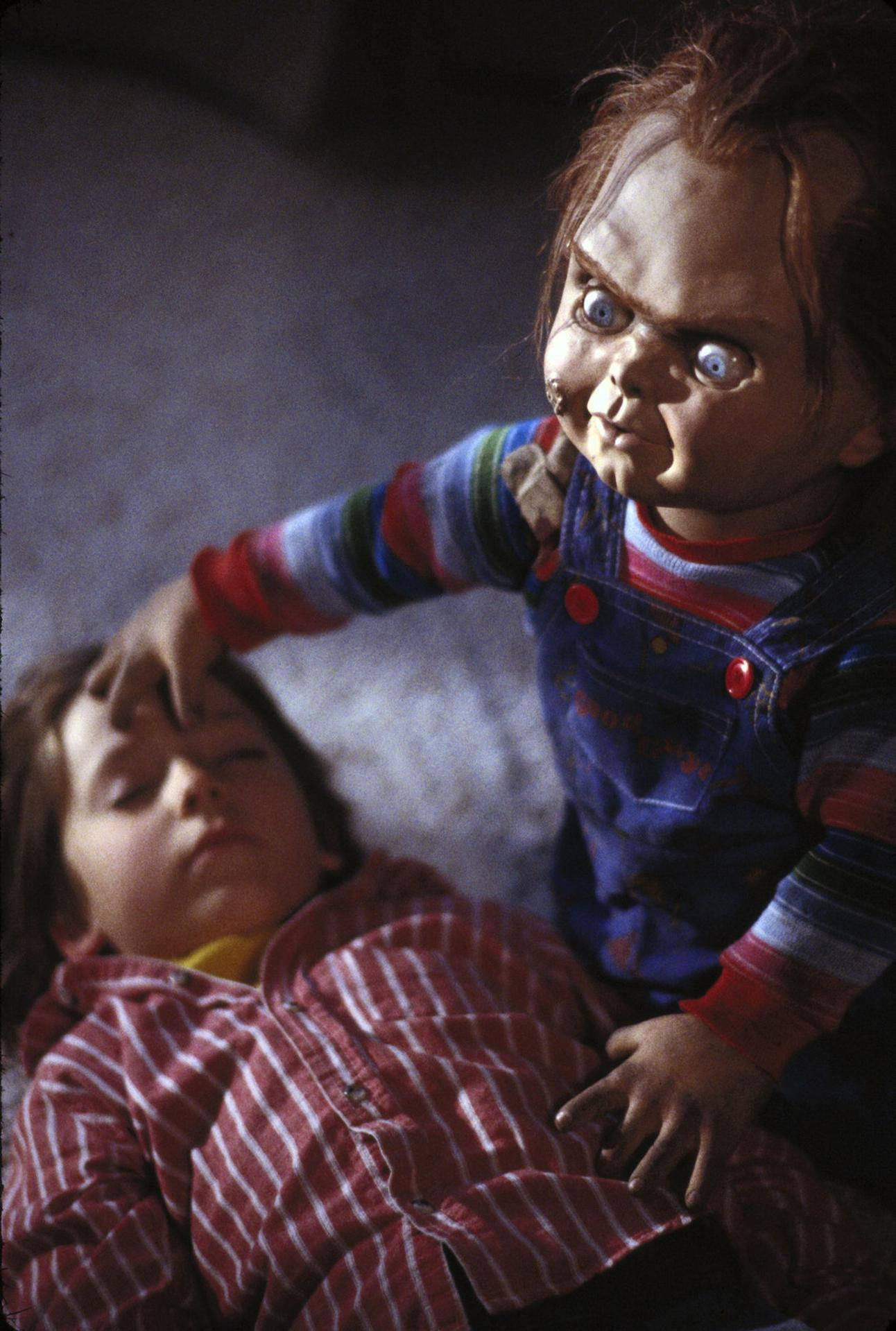 Eine Chucky-Puppe berührt den Kopf eines Kindes. Das Kind liegt. Die Puppe hat wütende Augen und trägt blaue Latzhosen.