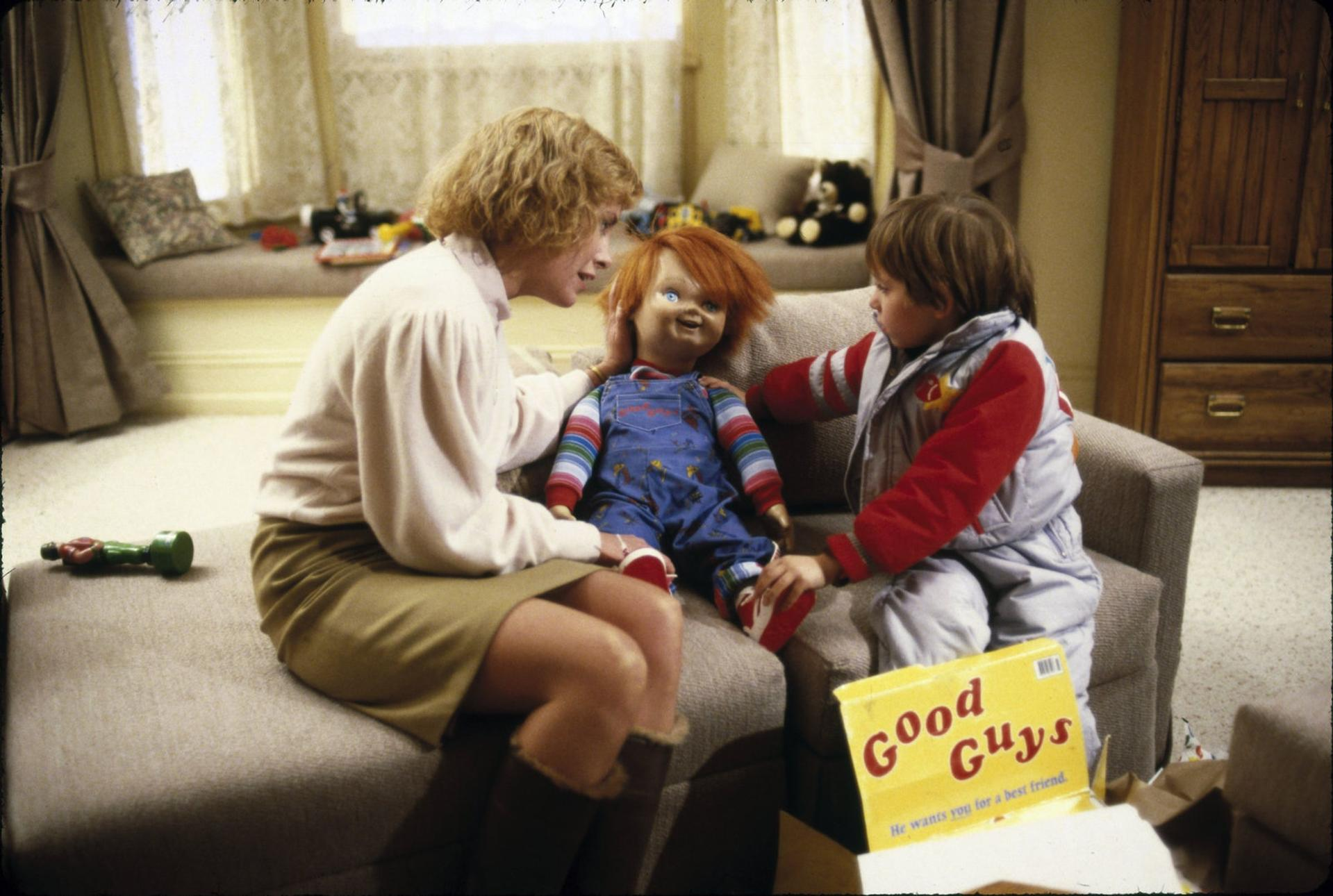 Eine Frau und ein Junge sitzen mit einer Puppe. Die Puppe ist Chucky. Eine Good Guys-Box ist geöffnet. Im Hintergrund sind ein Fenster und ein Holzschrank zu sehen.