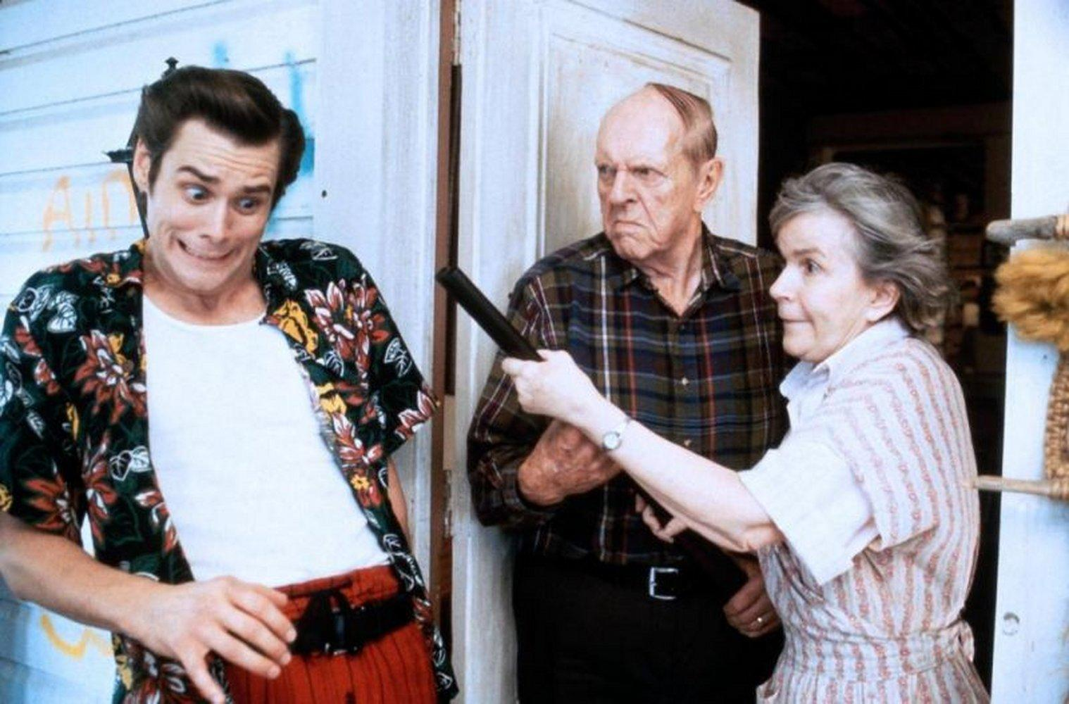Jim Carrey mit ängstlichem Gesichtsausdruck, ein Mann und eine Frau halten eine Schrotflinte. Szene aus einem Film, drinnen.