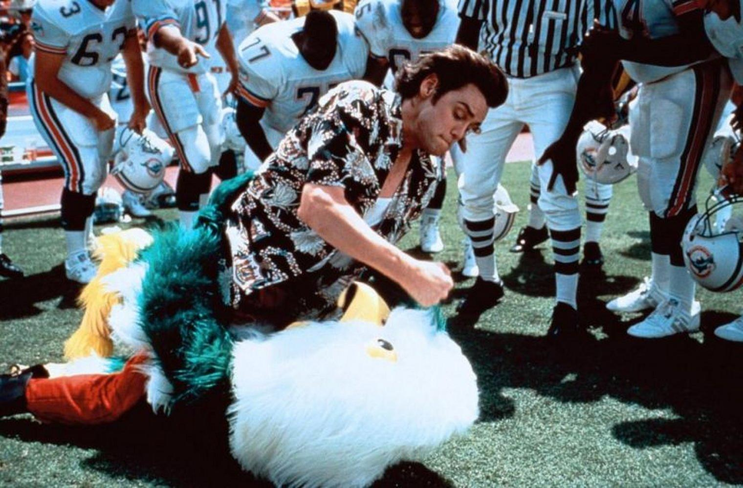 Ein Mann in einem Hawaiihemd kämpft mit einem Maskottchen auf einem Footballfeld, umgeben von Footballspielern und einem Schiedsrichter.
