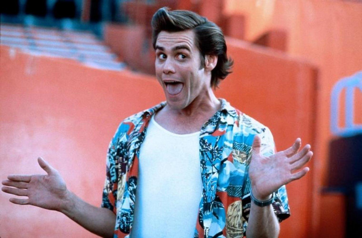 Jim Carrey mit offenem Mund und Armen. Er trägt ein buntes Hemd über einem weißen Hemd. Der Hintergrund ist orange.