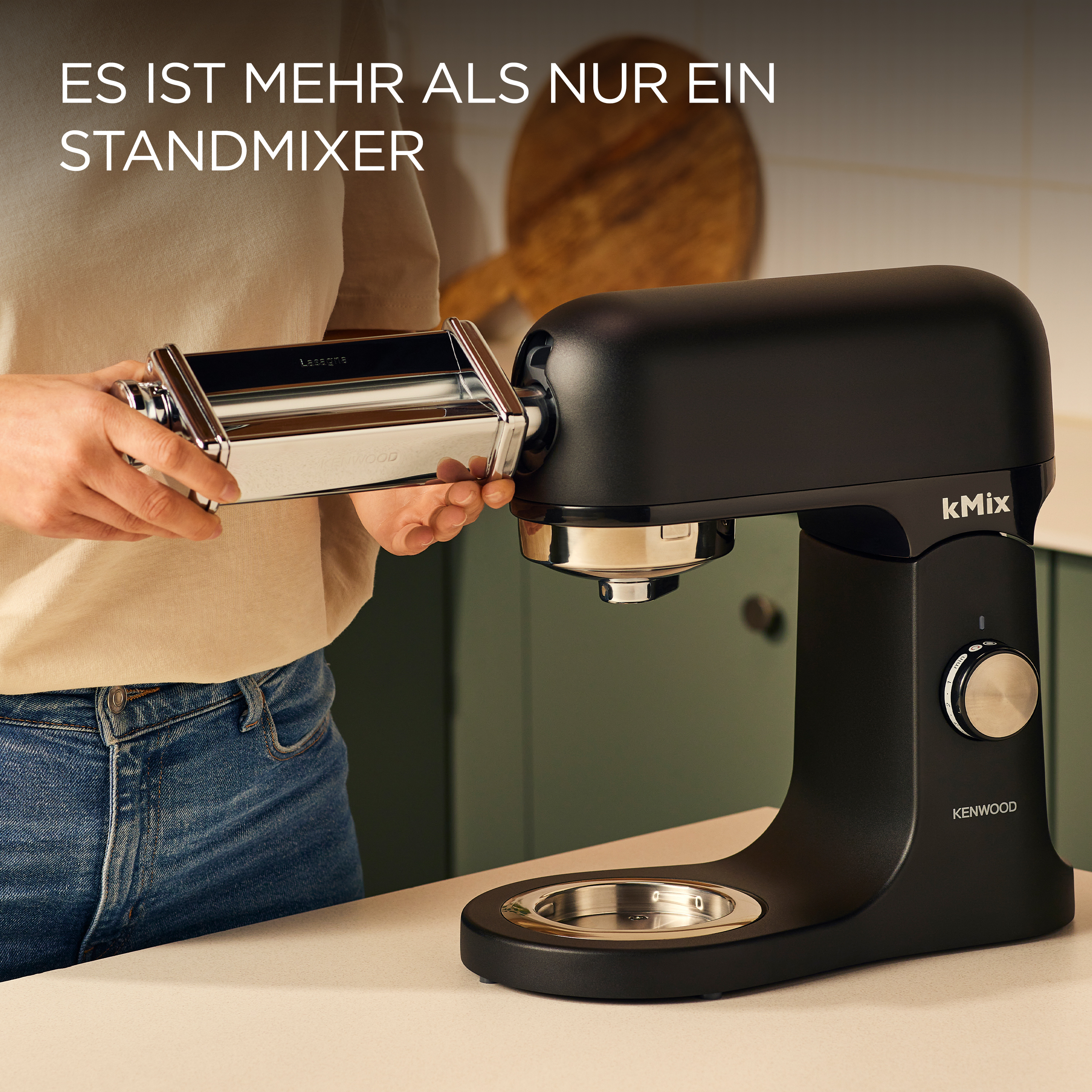 Ein schwarzer Kenwood kMix-Küchenmixer mit einem Nudelwalzenaufsatz. Eine Person befestigt die Walze. Grüner Hintergrund.