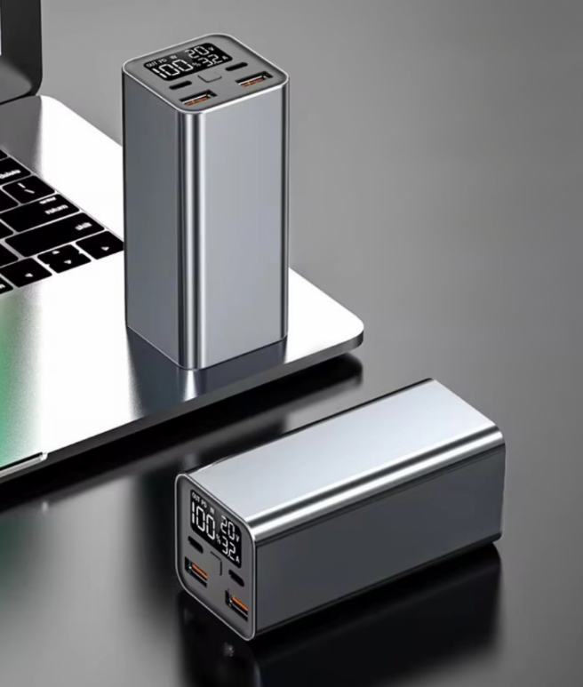 Ezüst power bank digitális kijelzővel és USB portokkal, laptopon és felületen. Egy másik power bank mellette.