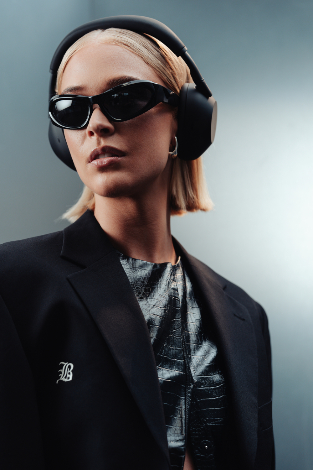 Een vrouw met zonnebril, koptelefoon, zwarte blazer en top met reptielenpatroon, blond haar, tegen een grijze achtergrond.