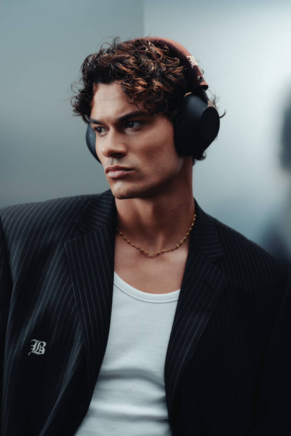 Een man met een koptelefoon, een blazer met krijtstrepen en een gouden ketting. Hij kijkt naar links.