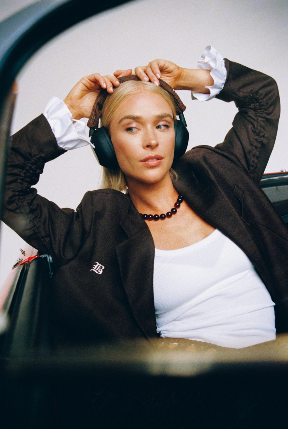 Blonde vrouw met koptelefoon en bruine blazer. Ze zit in een auto en kijkt weg.