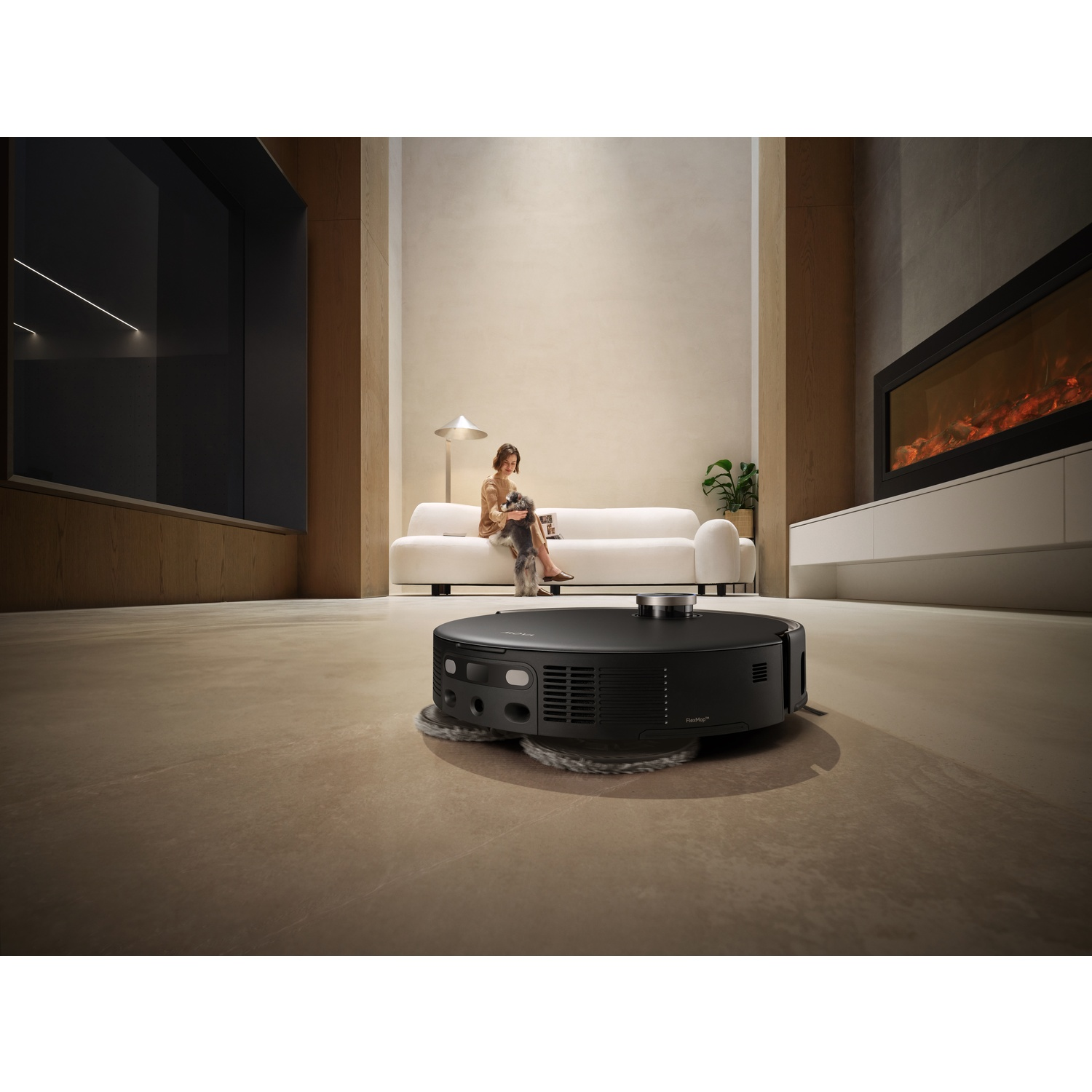 Un aspirateur robot noir nettoie une pièce. Une femme est assise sur un canapé blanc, avec un chien. Une cheminée est allumée. La pièce est moderne.