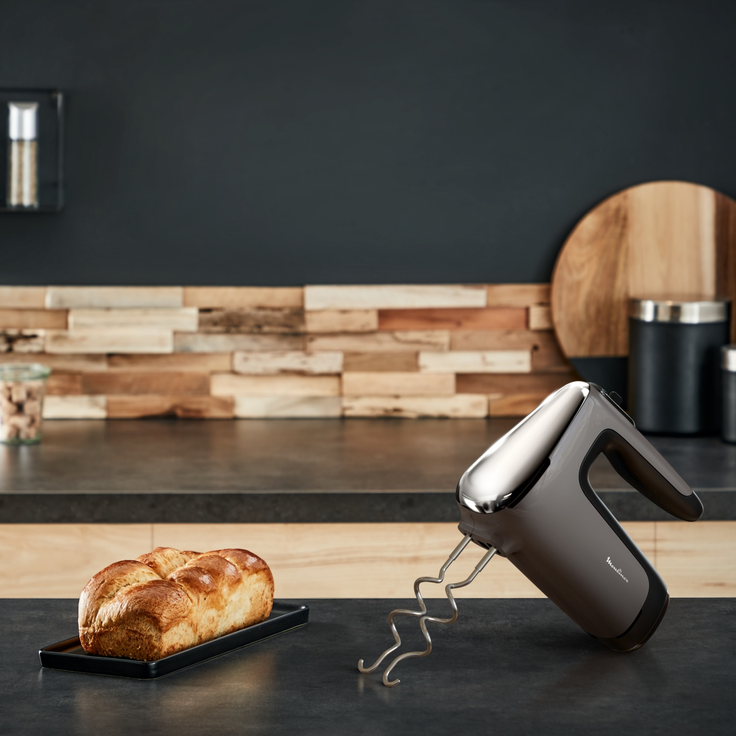 Un mixeur est posé sur un comptoir. Un pain est sur un plateau noir. Arrière-plan de cuisine avec du bois et des surfaces sombres.