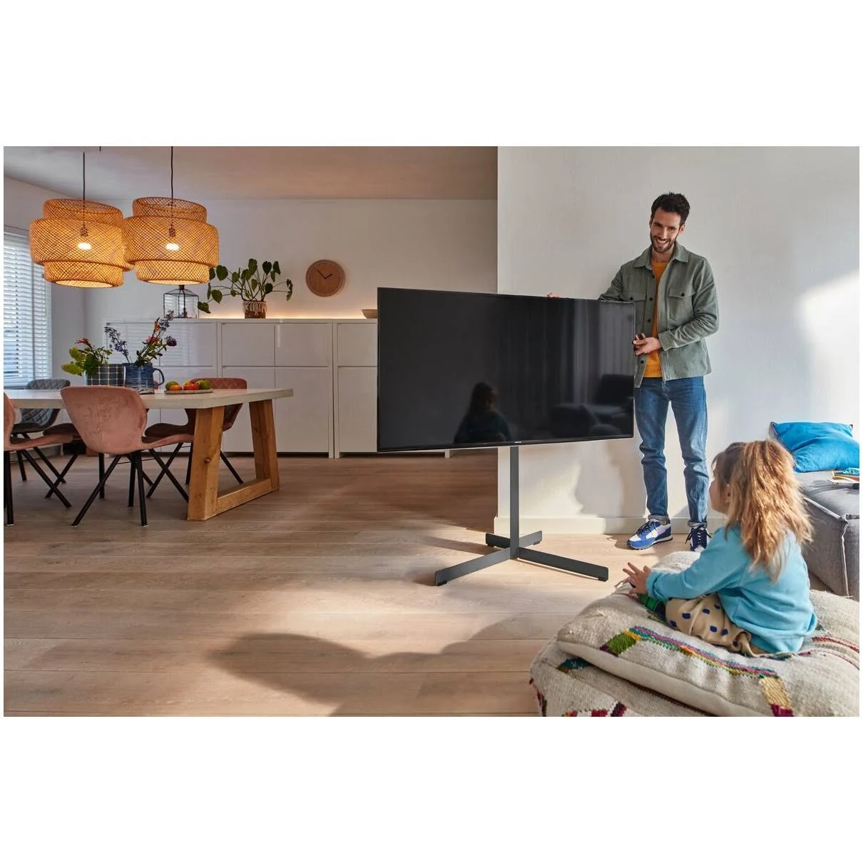Un hombre está de pie junto a un televisor grande, mostrándoselo a una niña sentada en un cojín. Se ve una mesa de comedor y lámparas colgantes.