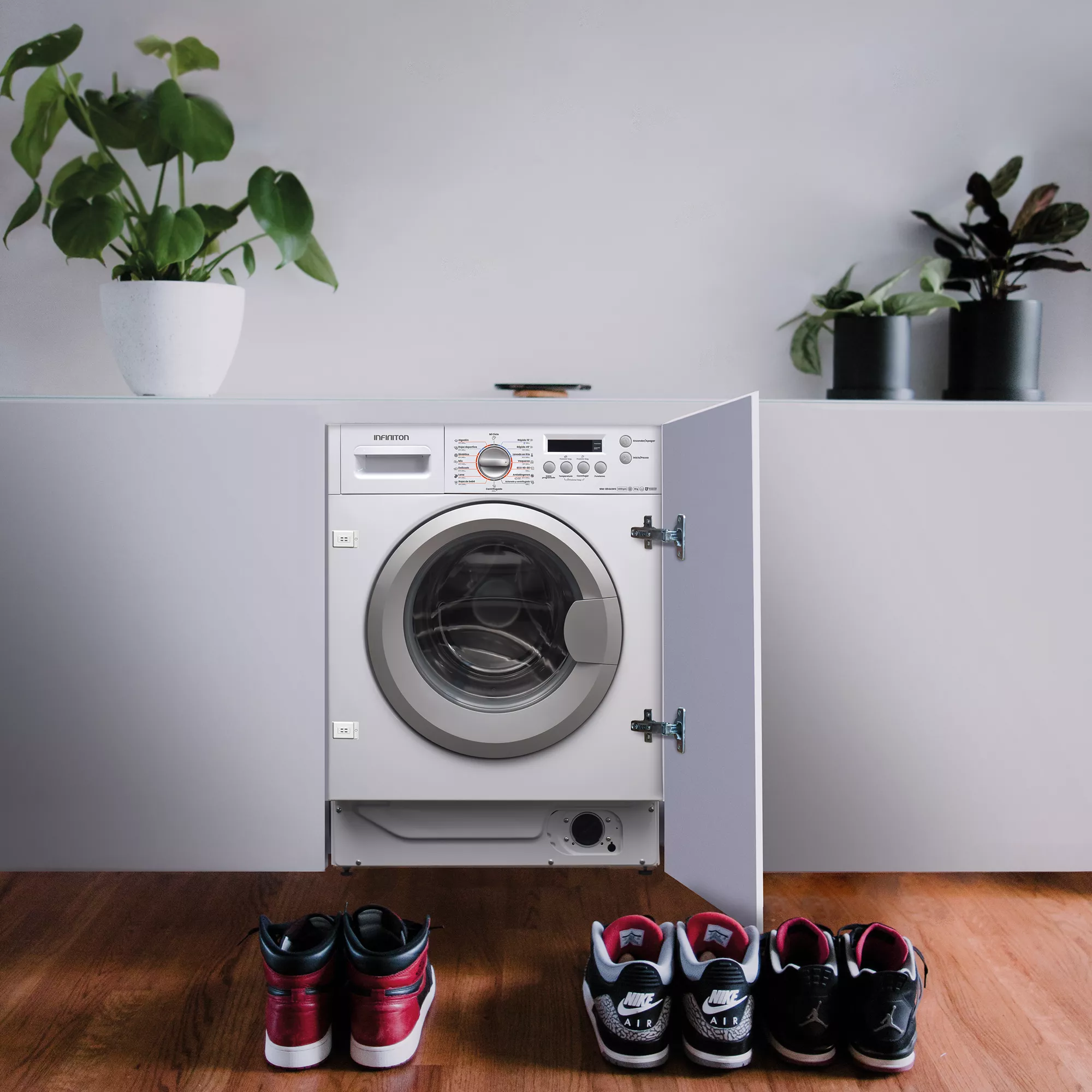 Una lavadora está en un armario, hay zapatos delante de ella, con plantas encima. La puerta está abierta.