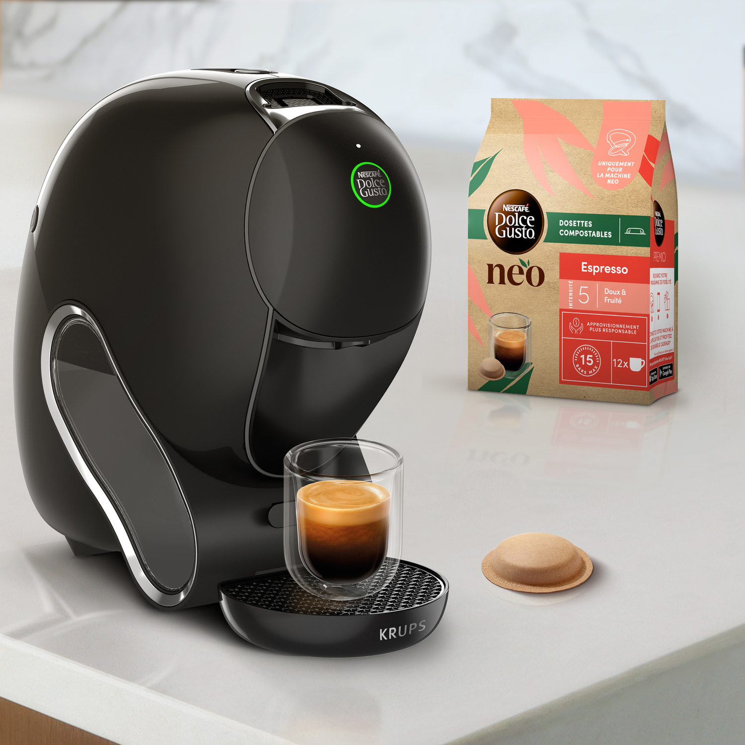 A black coffee machine with a glass of coffee. A coffee pod, and a coffee pod package are also present on the table.