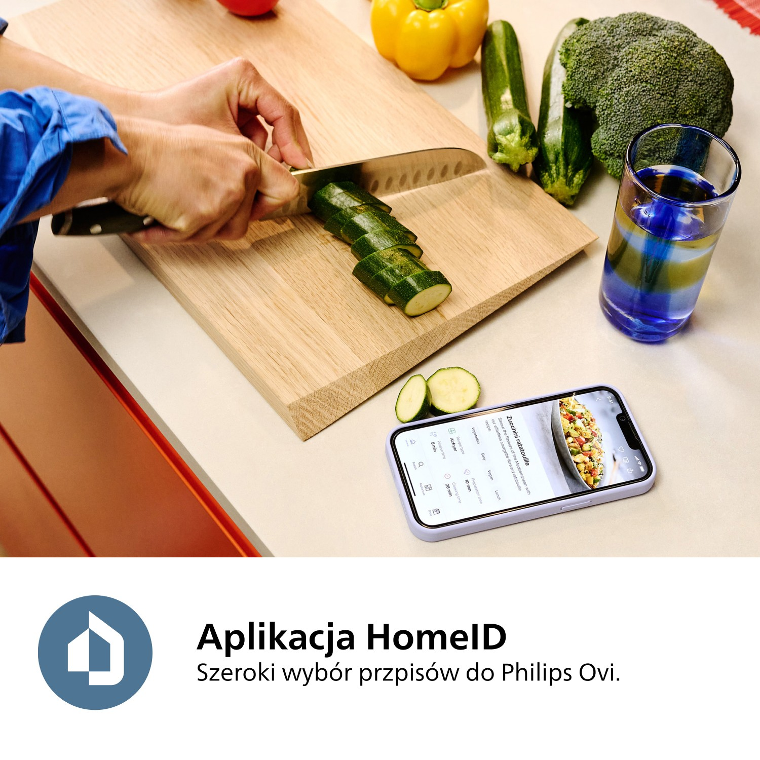 Osoba kroi cukinię na drewnianej desce. Warzywa, takie jak brokuły i żółta papryka, są w pobliżu, z telefonem pokazującym przepis.