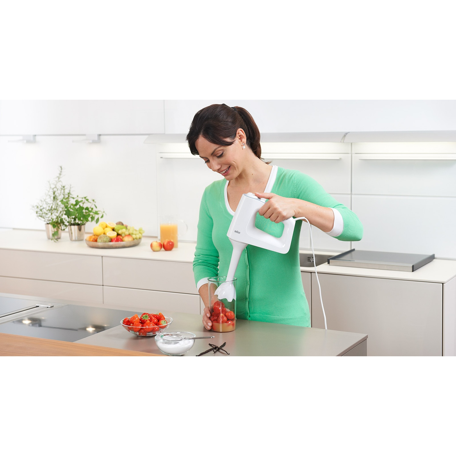 Vrouw die aardbeien mixt in een keuken met een staafmixer. Andere ingrediënten en fruit op het aanrecht.