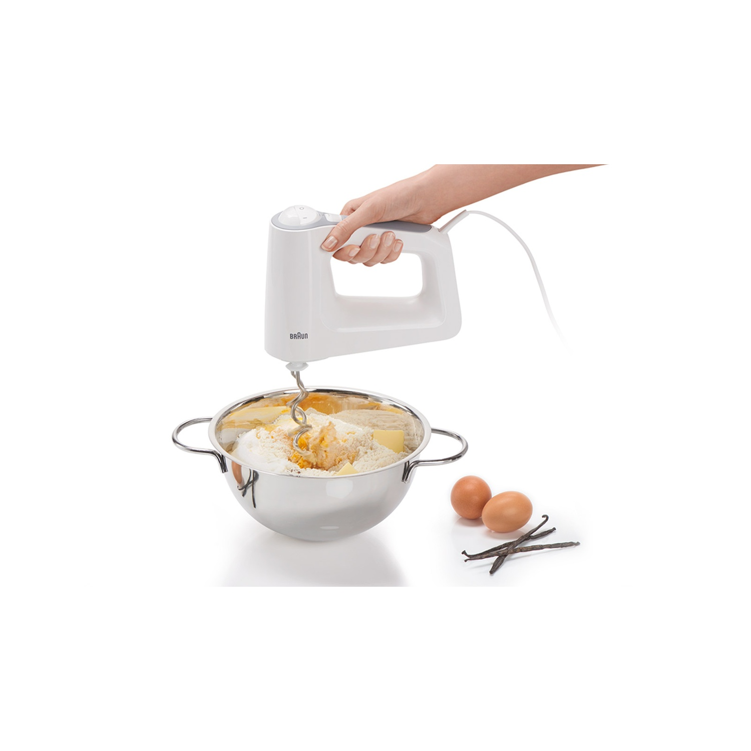 Een hand houdt een witte Braun mixer vast, ingrediënten mixend in een roestvrijstalen kom. Eieren, vanillestokjes ernaast.