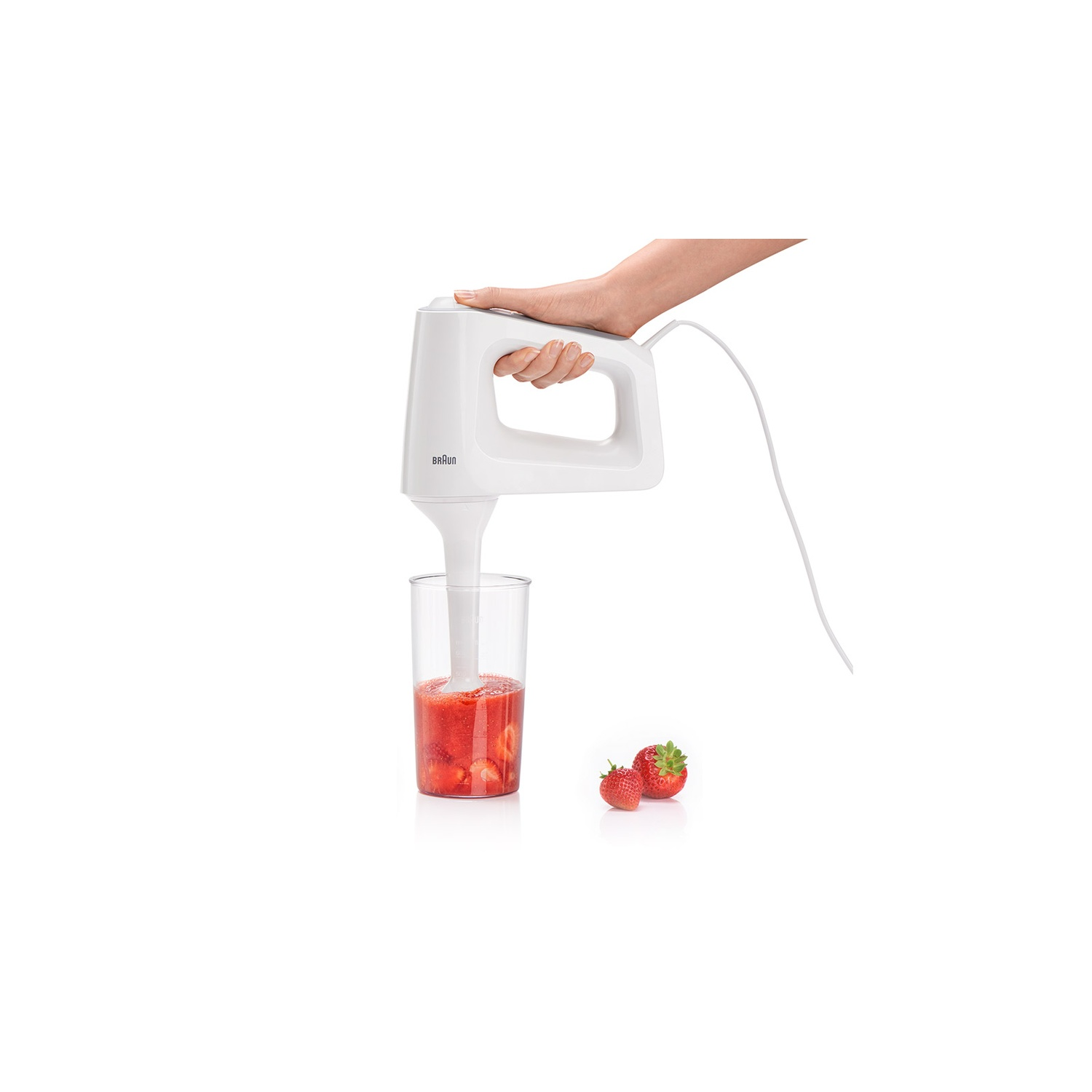 Een hand houdt een witte blender vast, die aardbeien in een glas mixt. Twee aardbeien staan rechts.