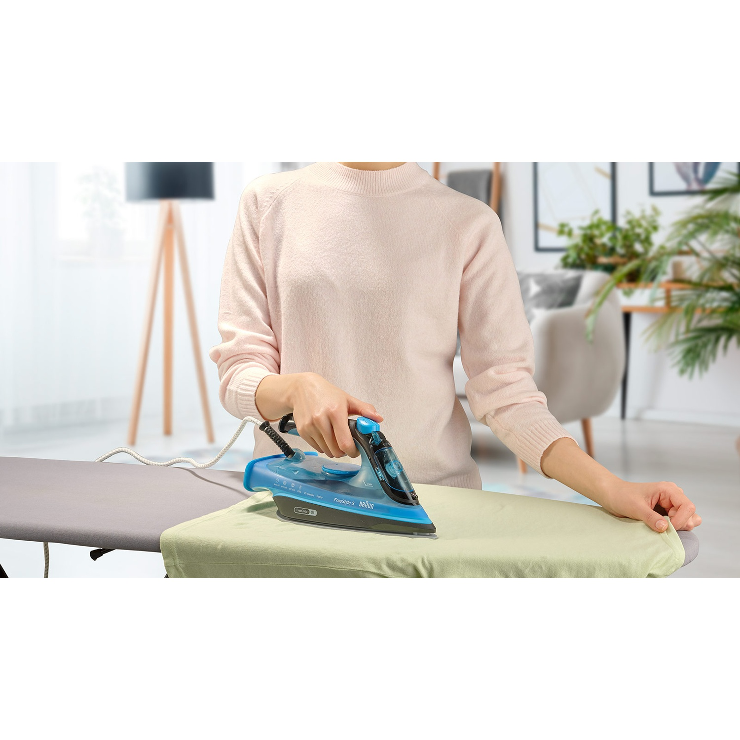 Une personne repasse un vêtement sur une planche à repasser. Elle tient un fer à repasser bleu sur le vêtement avec un pull rose clair.