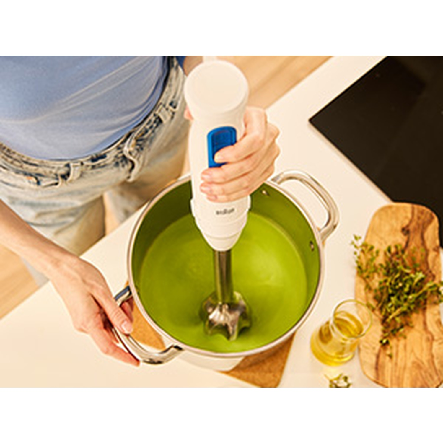 A person is using a hand blender in a pot of green soup. Herbs, oil, and a cutting board are nearby.