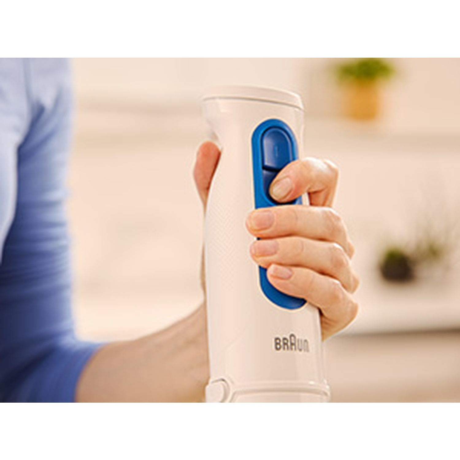 A person holds a white Braun hand blender. The hand presses a blue button on the blender. A blue shirt is visible.