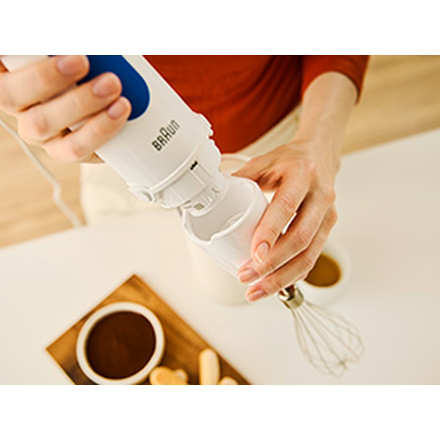 A person using a Braun hand blender. The blender is white and blue. There are bowls with brown liquid and cookies on a wooden board.