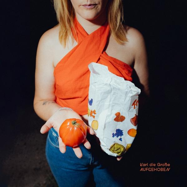 Eine Frau hält eine Tomate und eine Papiertüte mit Essensillustrationen. Sie trägt ein orangefarbenes Oberteil und blaue Jeans. Dunkler Hintergrund.
