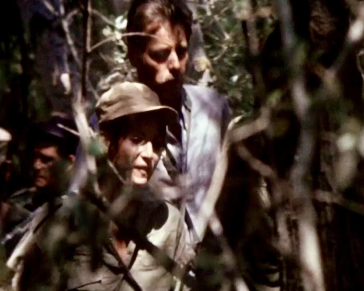 Drei Personen in einem Wald. Einer trägt eine Kappe, zwei andere sind im Hintergrund. Bäume und Blätter verdecken Teile des Bildes.
