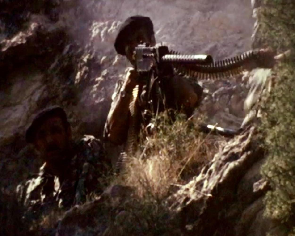 Zwei Soldaten in felsigem Gelände, einer bedient ein Maschinengewehr. Sie tragen dunkle Baretts und Tarnkleidung. Vegetation und Felsen sind sichtbar.