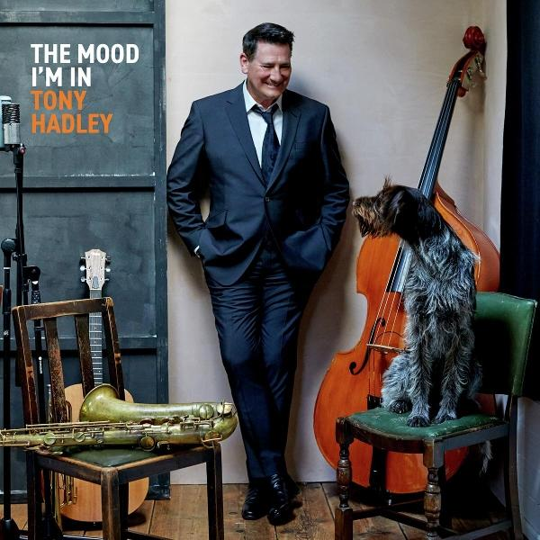 Mann im Anzug lehnt an einer Wand, Hund sitzt auf einem Stuhl. Instrumente und Stühle sind im Vordergrund, Albumtitel links.