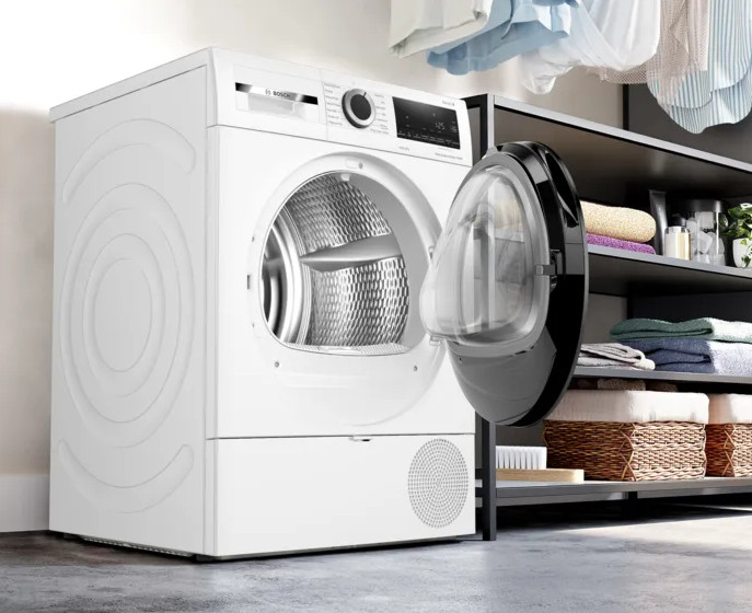 Biała suszarka z otwartymi drzwiami, odsłaniająca bęben. Półki z ręcznikami i koszami w tle. Ubrania wiszą nad.