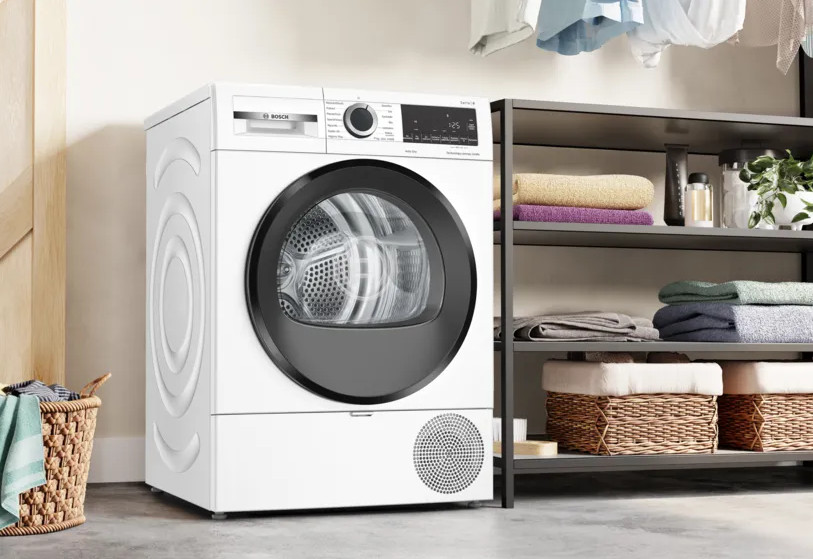 Biała suszarka Bosch stoi obok półki z ręcznikami i koszami. Pranie wisi nad głową. Kosz ubrań na pierwszym planie.
