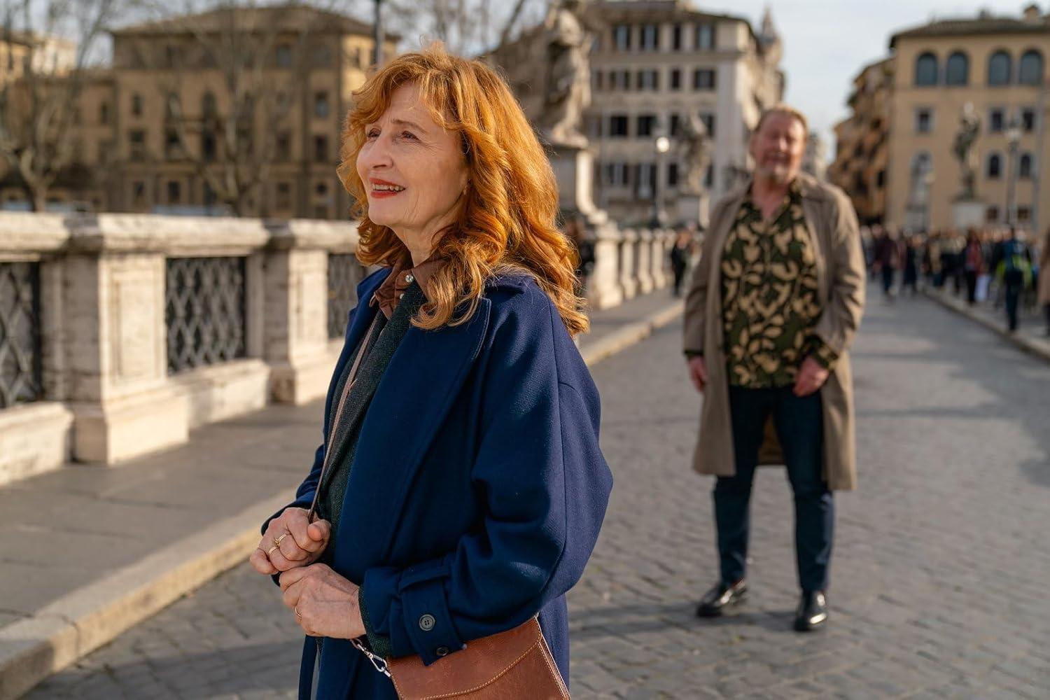 Frau mit roten Haaren in blauem Mantel auf einer Brücke. Mann im Hemd mit Blumenmuster und Mantel im Hintergrund. Gebäude und Menschen sind sichtbar.