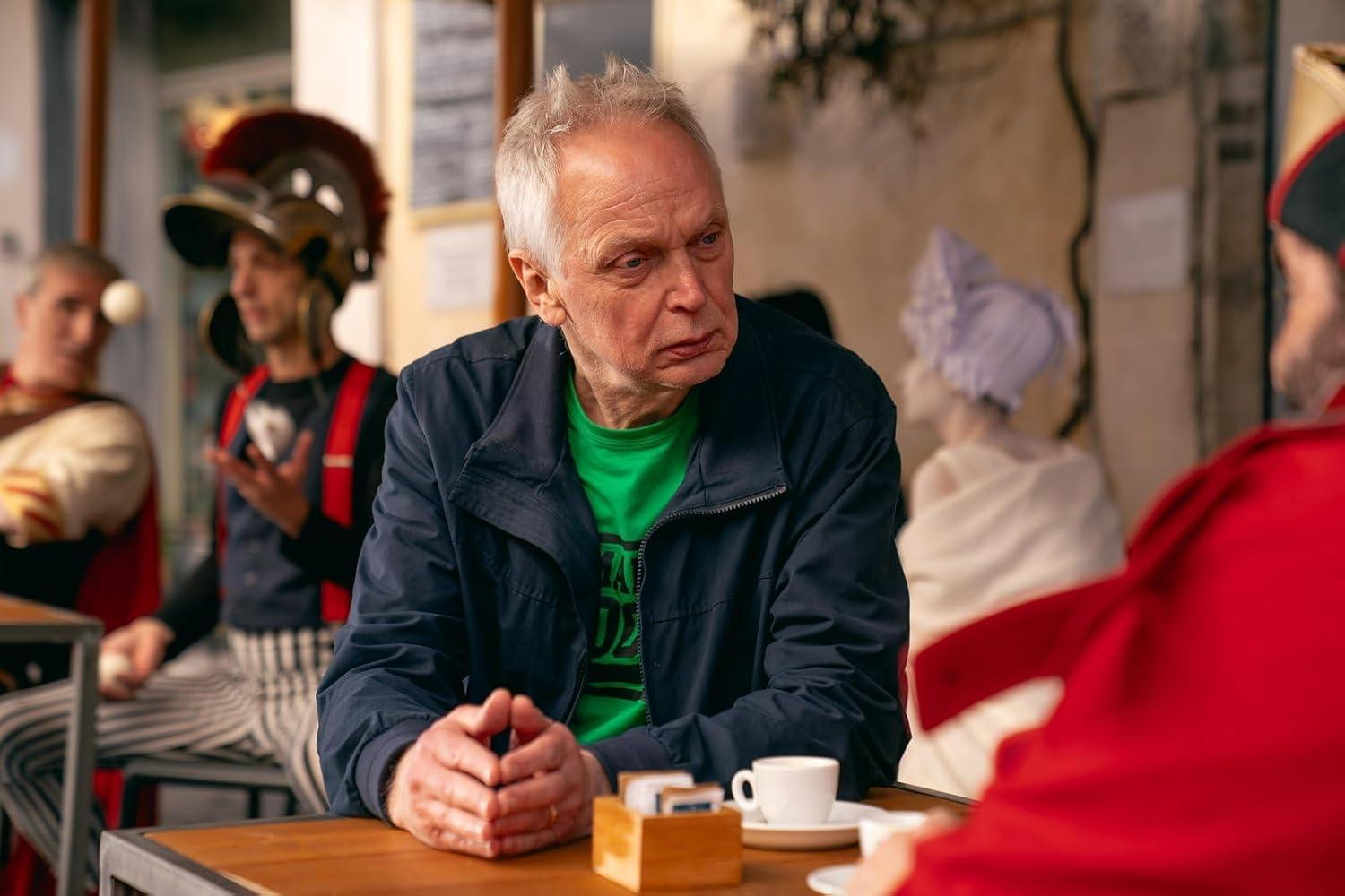 Ein älterer Mann in grünem Hemd und blauer Jacke sitzt an einem Tisch. Andere in Kostümen sind im Hintergrund.