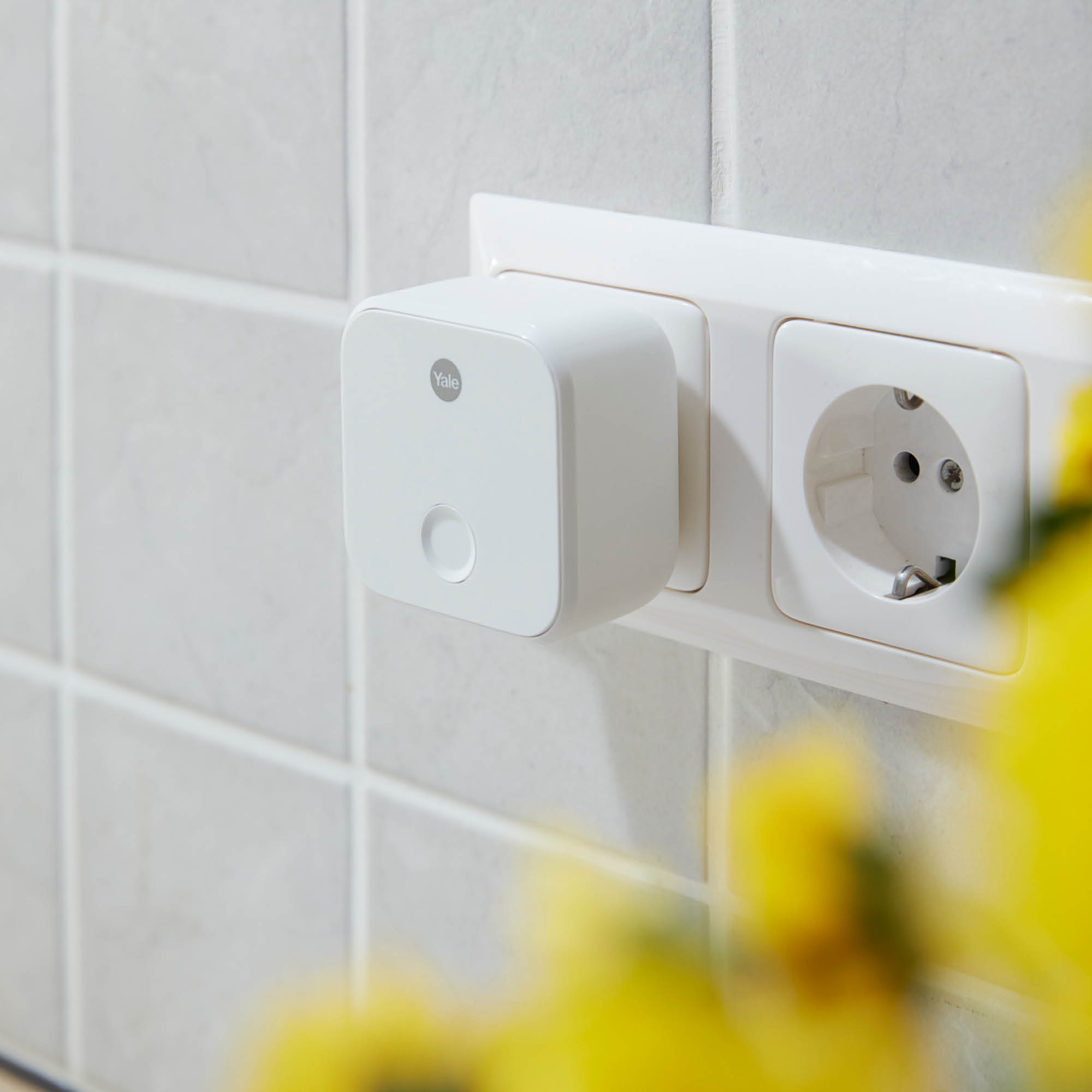 Ein weißer Yale Smart Plug ist in eine Steckdose eingesteckt. Gelbe Blumen sind im Vordergrund und weiße Fliesen im Hintergrund.