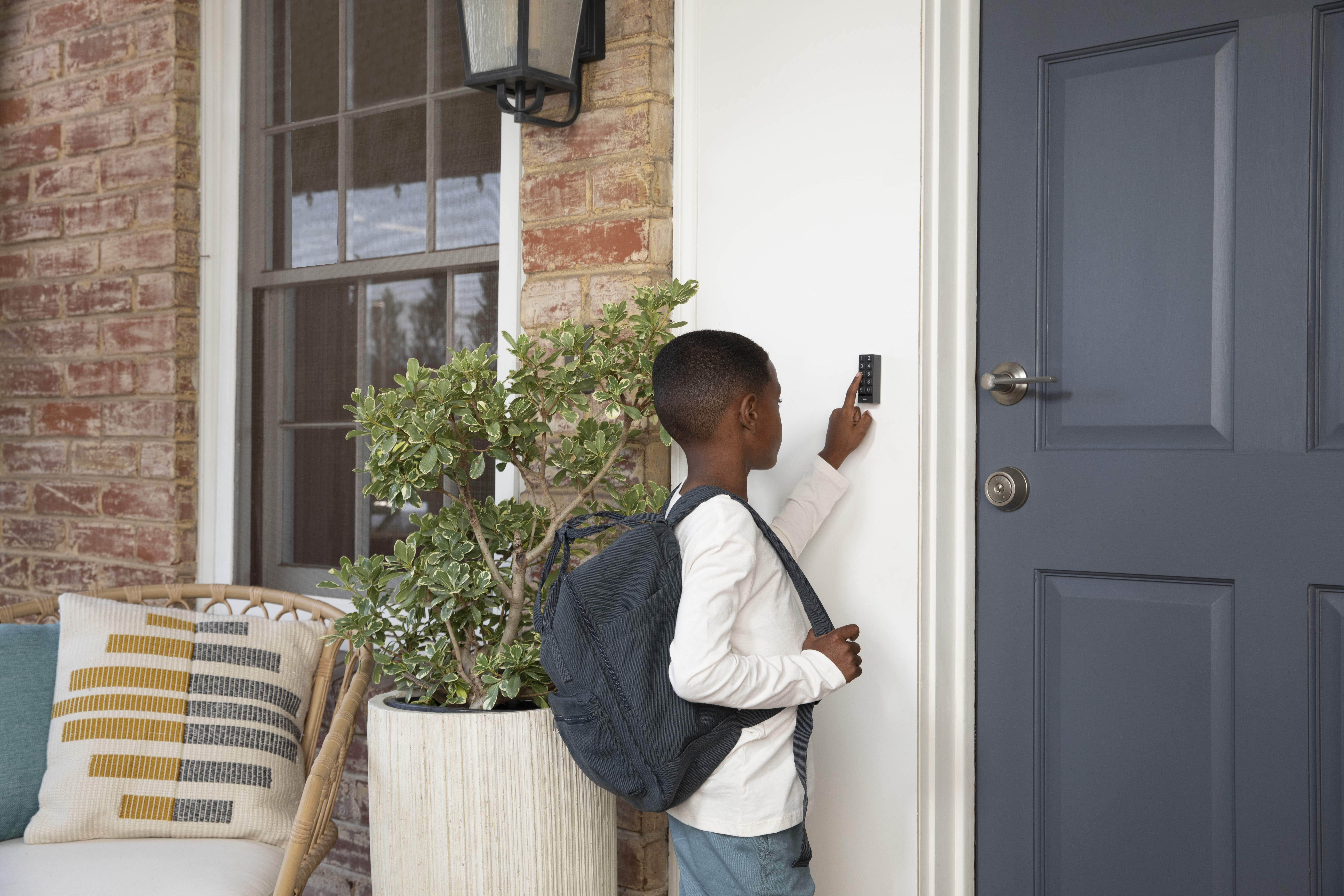 Ein Kind mit Rucksack betritt ein Haus und benutzt ein Tastenfeld an einer weißen Tür. Eine Ziegelmauer und eine Topfpflanze sind ebenfalls sichtbar.