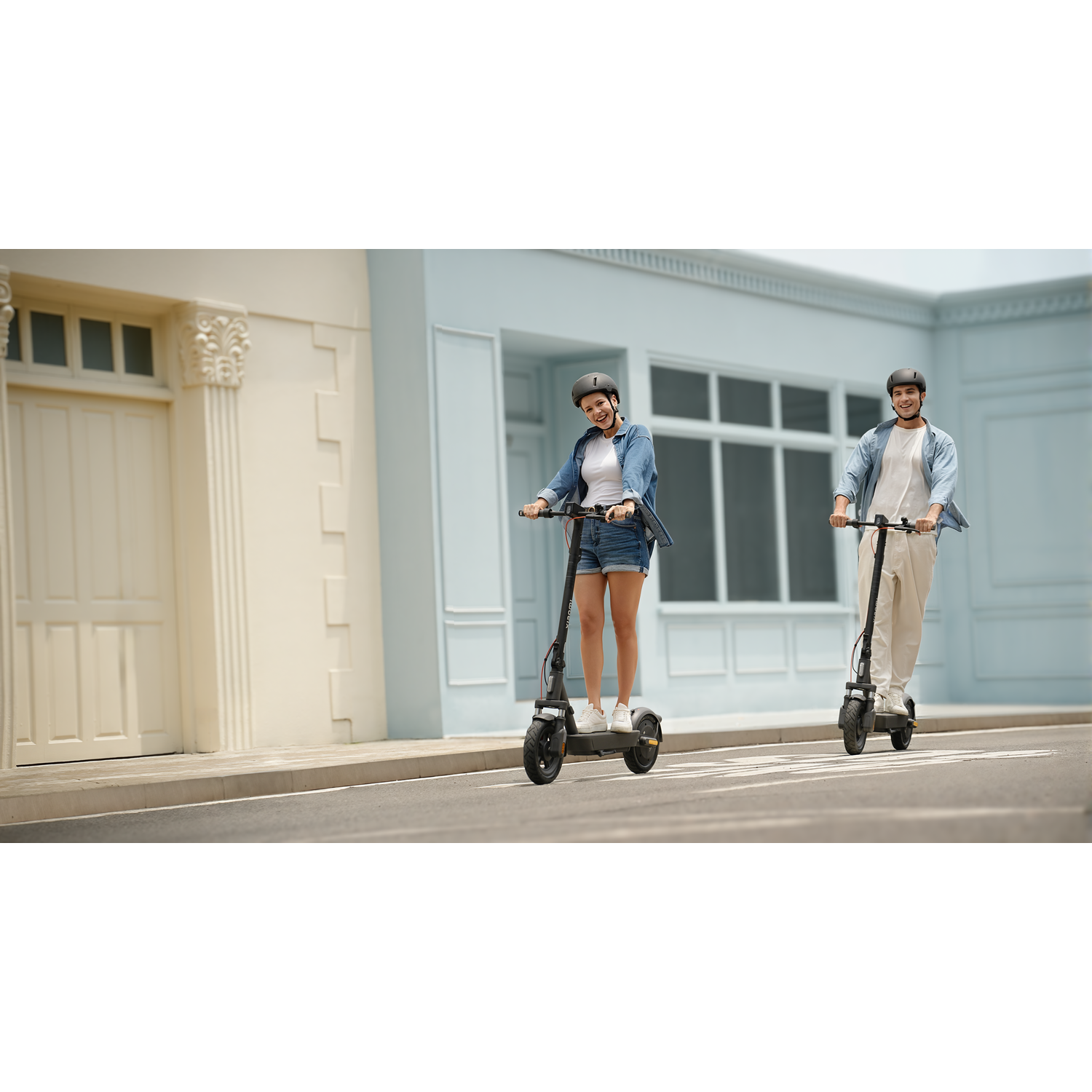 Two people on scooters, wearing helmets, smile at the camera. They ride on a street with buildings in the background. One is female.