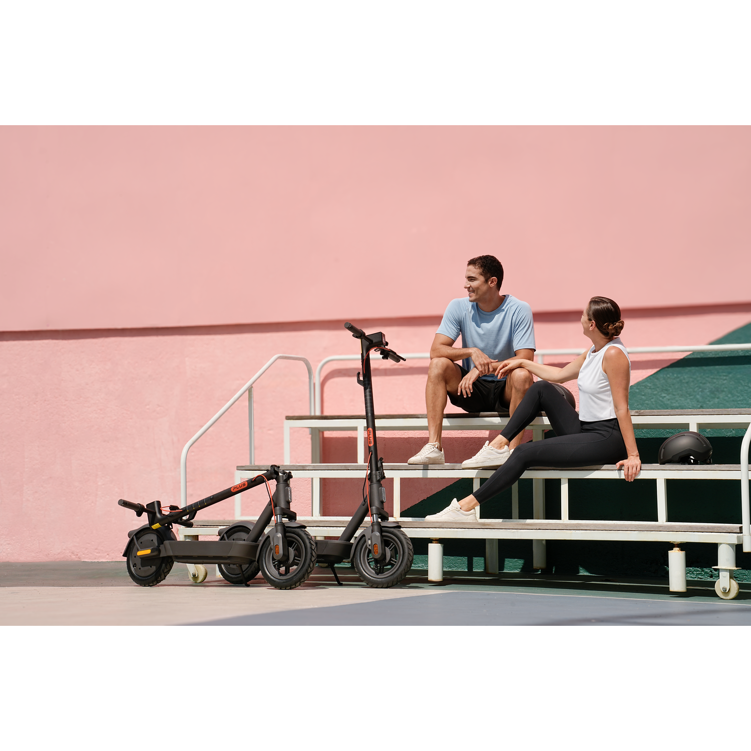 A man and woman sit on steps. Two electric scooters are nearby. A pink wall is in the background.