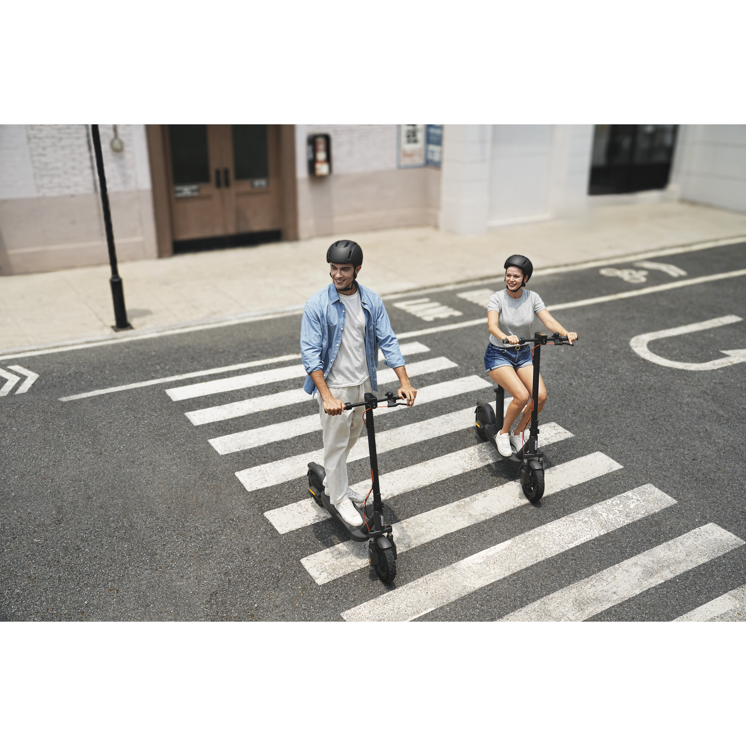 Two people on electric scooters on a crosswalk. One man wears a blue shirt, the woman wears a gray shirt. Both have helmets.