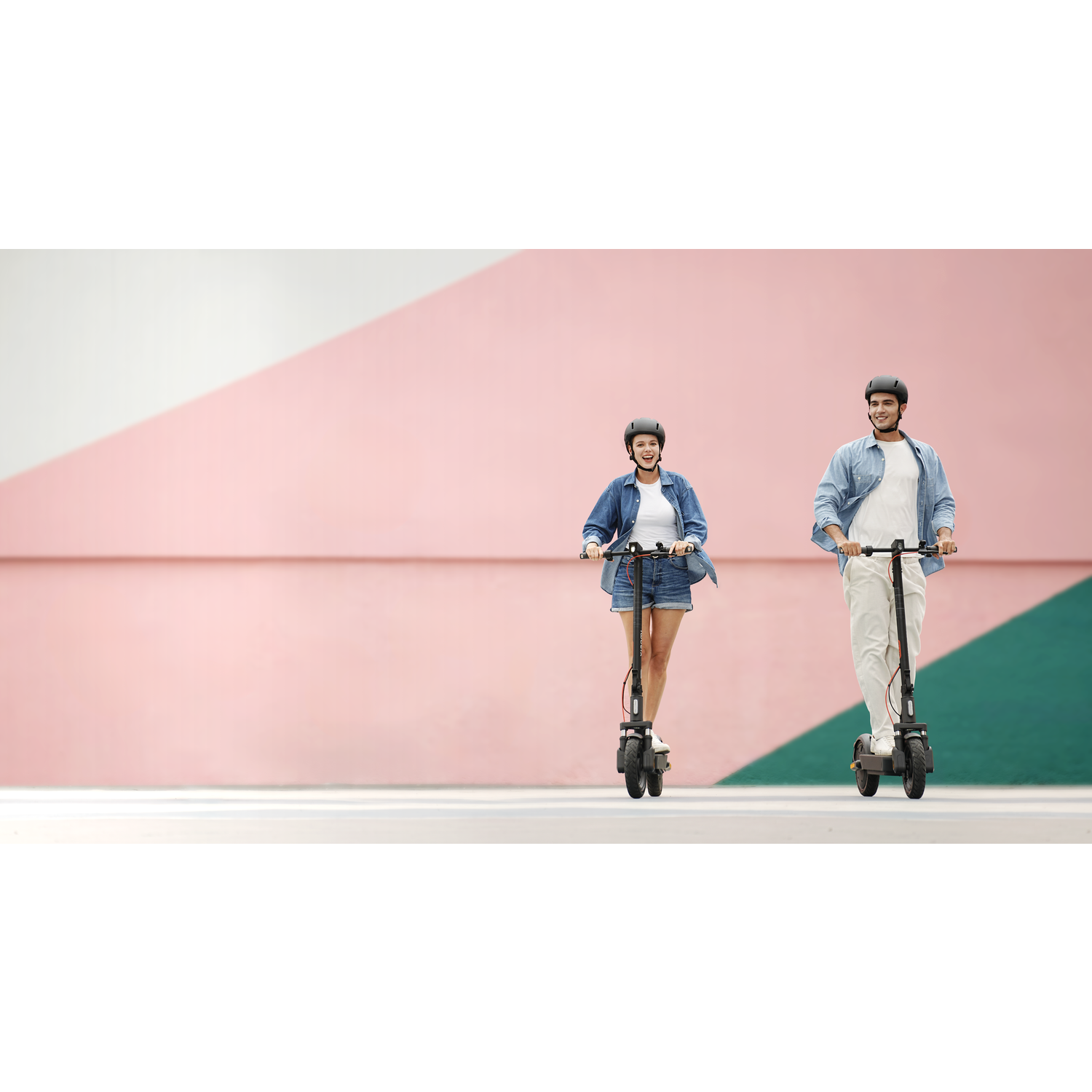 Two people on scooters, wearing helmets and casual clothing, against a pink and green wall. They are smiling and enjoying the ride.