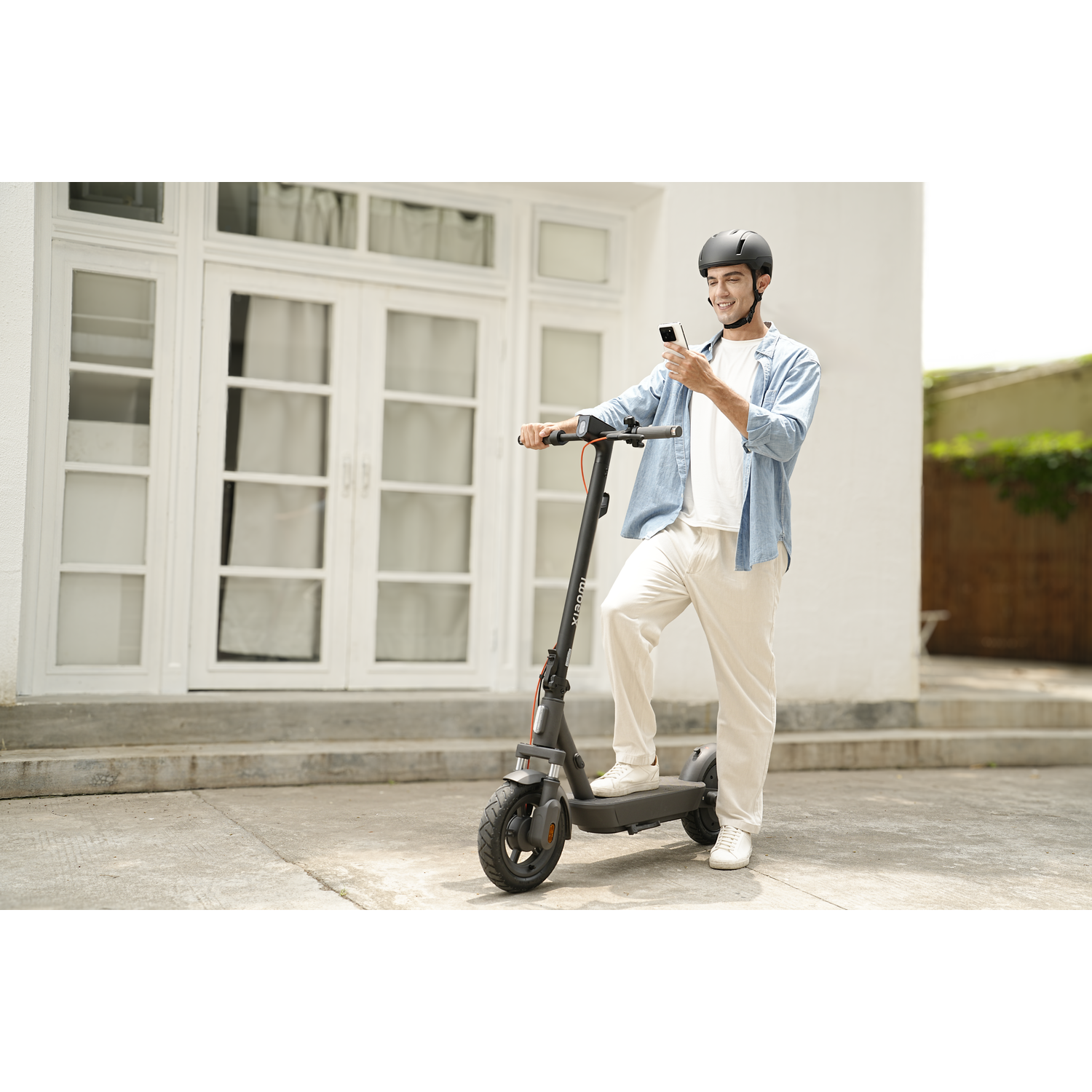 A man wearing a helmet stands with an e-scooter, holding a phone. He's smiling. Behind him is a white building and trees.