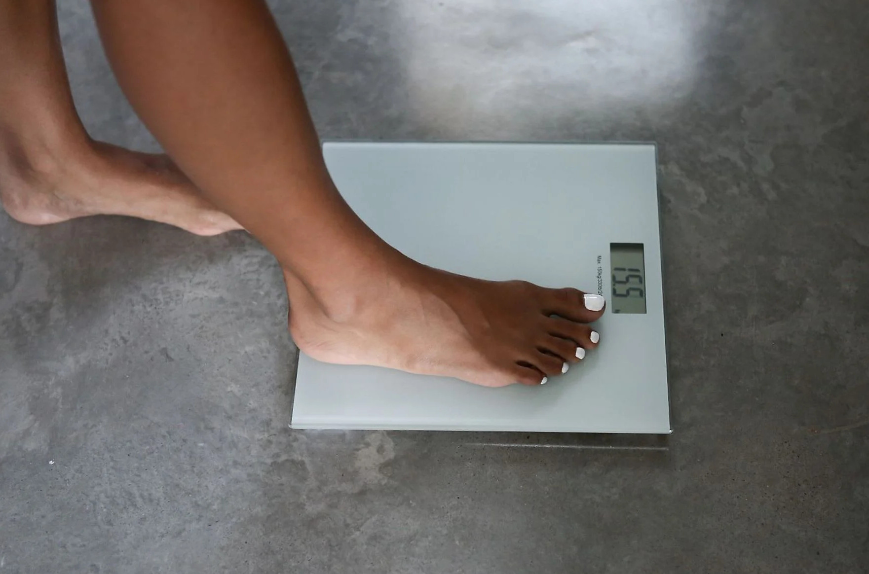 Bosa stopa osoby na wadze. Waga pokazuje 155,5. Paznokcie u stóp są pomalowane na biało. Podłoga jest szara.