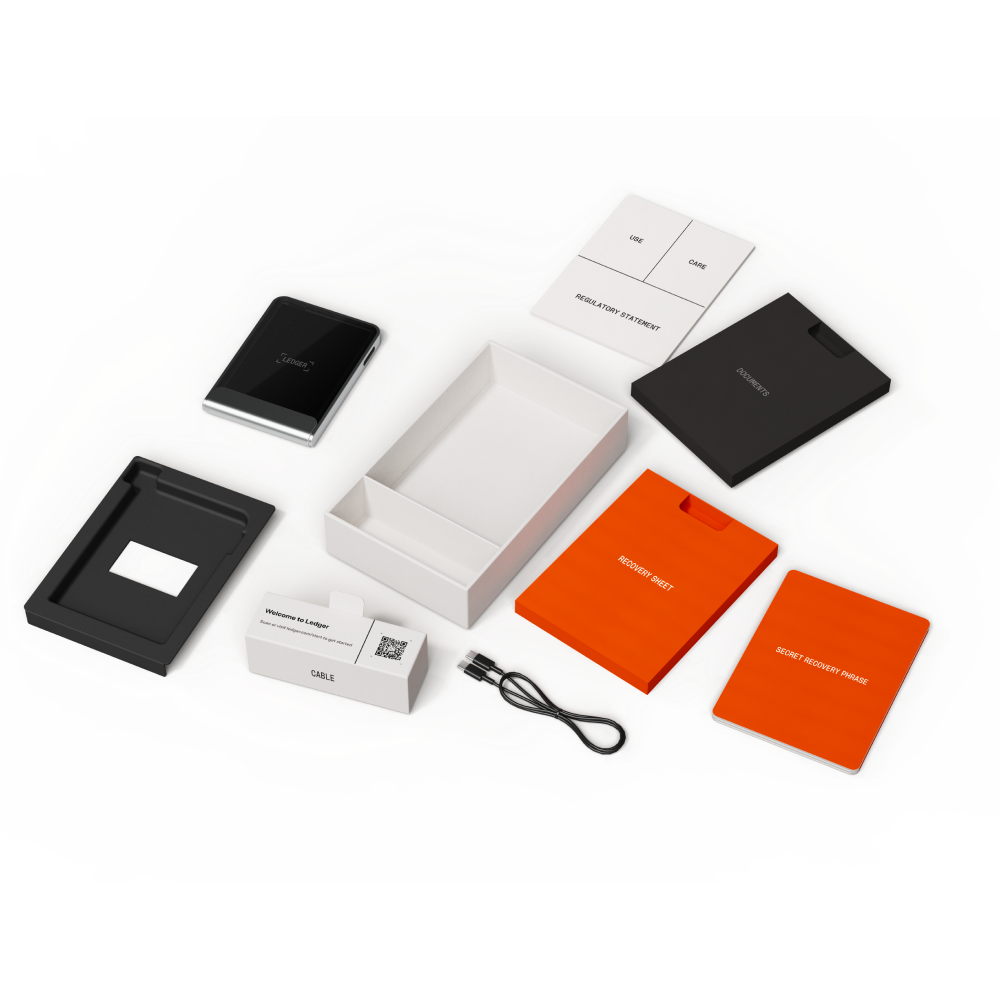 Kit de monedero hardware Ledger con dispositivo, documentación, cable y hojas de recuperación. Componentes sobre superficie gris.