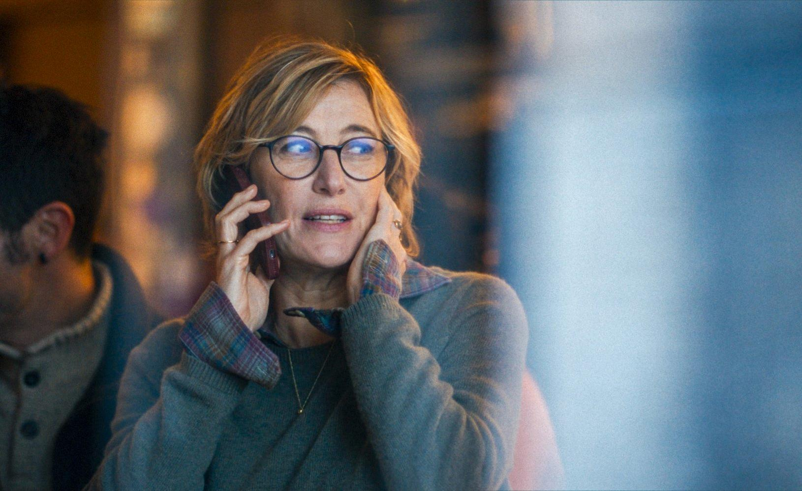 Eine Frau mit Brille telefoniert, trägt einen grauen Pullover und ein kariertes Hemd. Eine andere Person ist im Hintergrund unscharf.