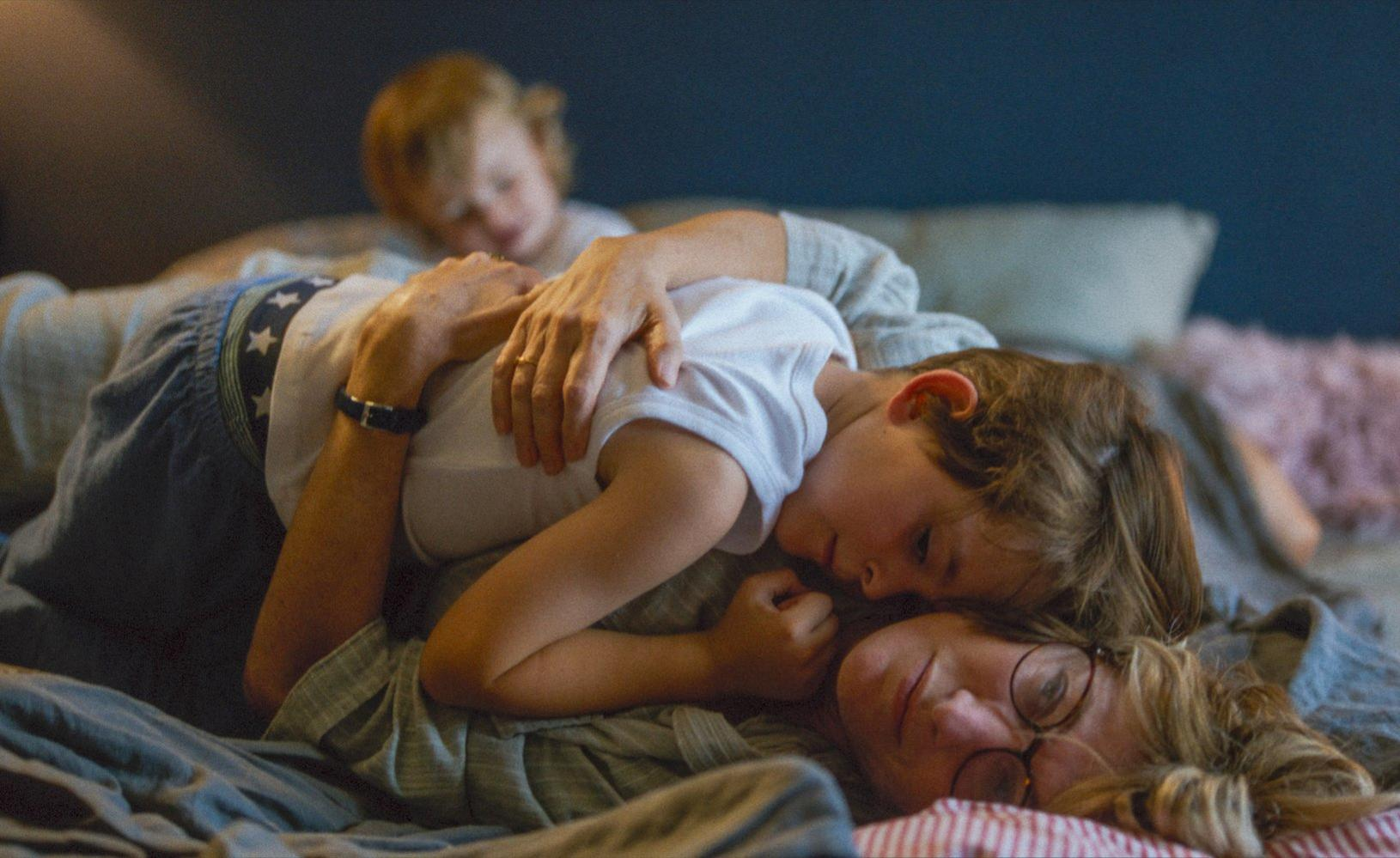 Eine Frau und zwei Kinder liegen auf einem Bett. Die Frau trägt eine Brille. Die Kinder tragen weiße Hemden.
